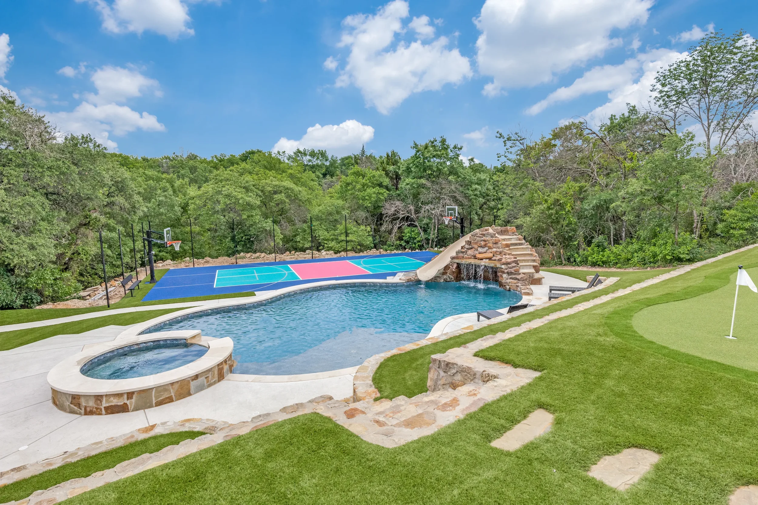 Backyard with swimming pool, hot tub, and a waterfall slide surrounded by grass, trees, and a blue sky with clouds.