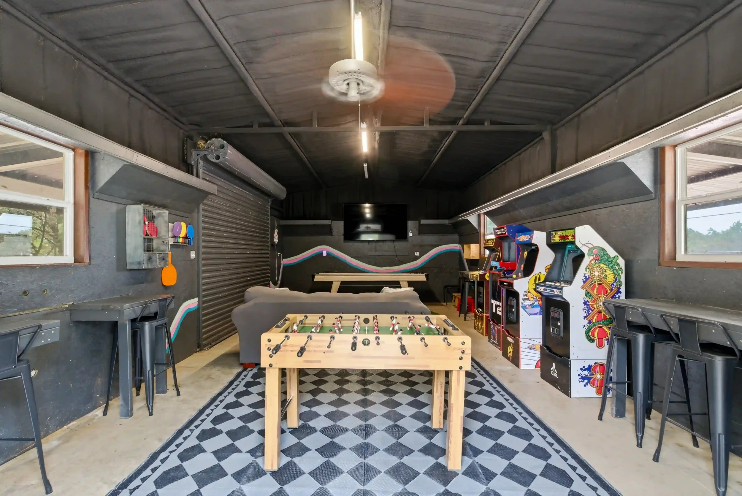 A game room with arcade machines on the right side, a foosball table in the center, a foosball table on the left, large windows on each side, and a wall-mounted TV at the back.