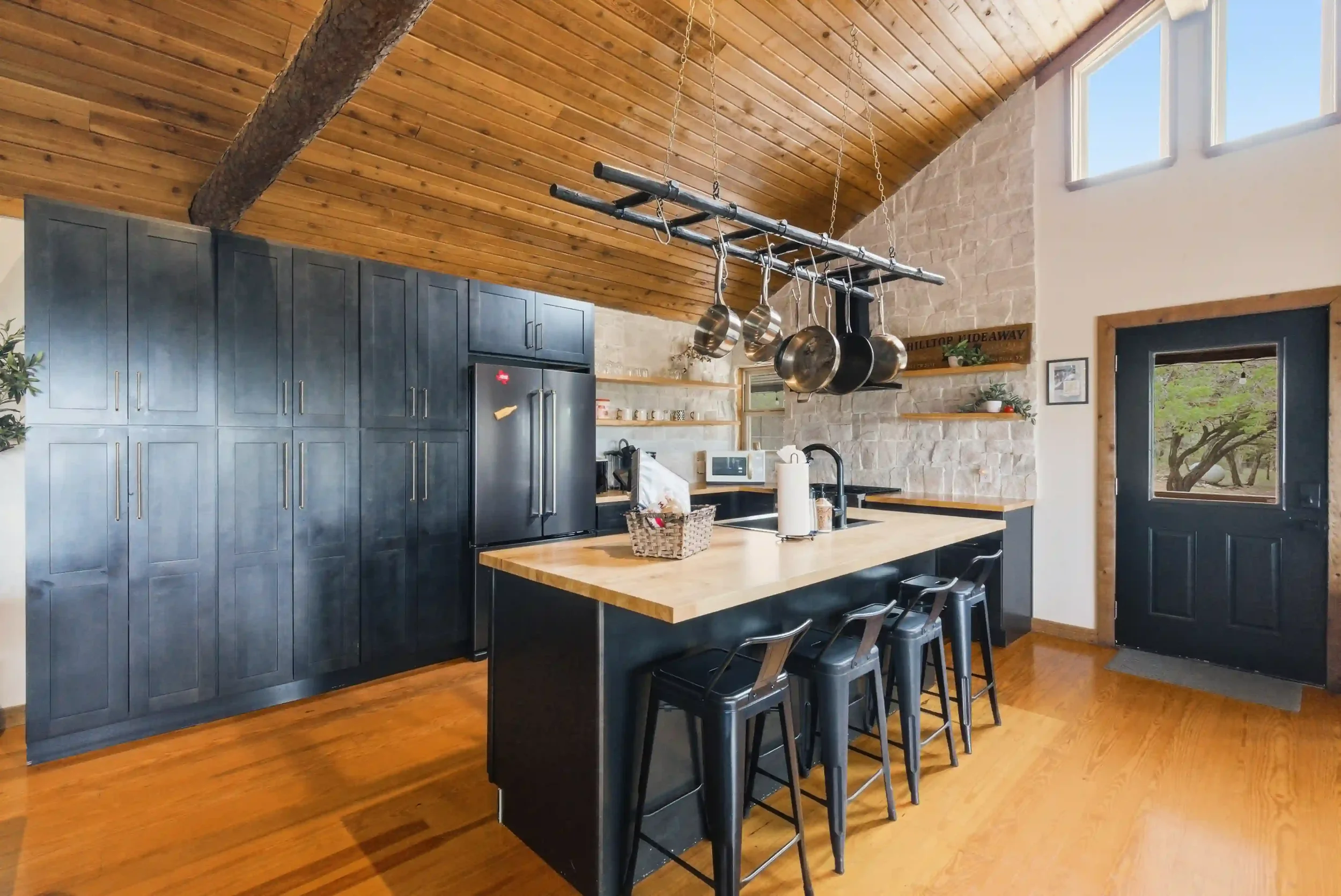 Modern kitchen with high vaulted wooden ceiling, black cabinets, stone wall, and large windows, with a kitchen island and hanging pots and pans.