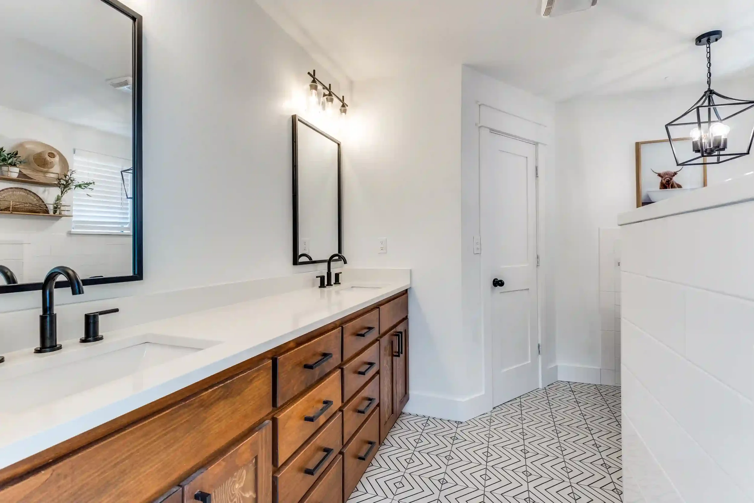 Modern bathroom with white walls, wooden vanity with black handles, black farmhouse sink faucet, large mirror, geometric patterned floor tiles, wall-mounted light fixtures, and a hanging geometric pendant light.