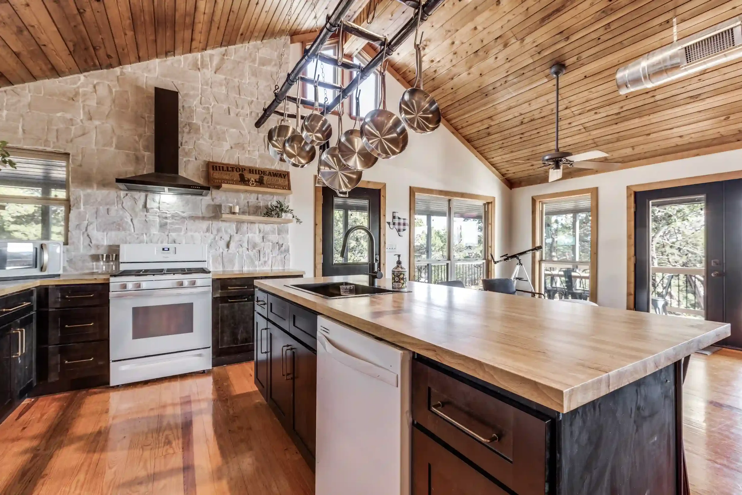 Kitchen with wooden ceiling, stone wall, hanging pots, white and dark cabinetry, island, and large windows.