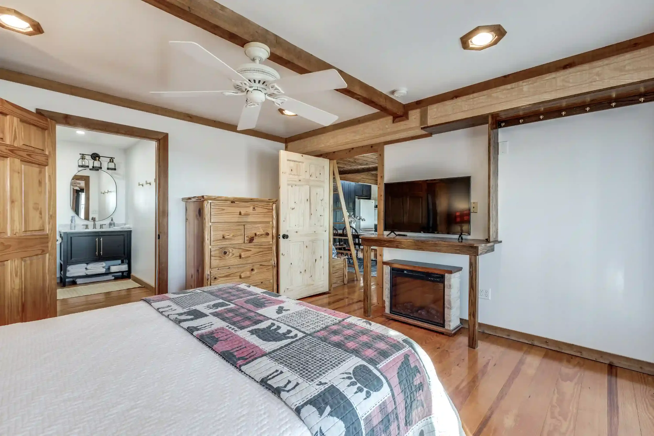 Interior view of a bedroom with a bed, wooden beams on the ceiling, a ceiling fan, a rough-cut wood divider, a TV, and an open doorway leading to a bathroom with a mirror and sink.