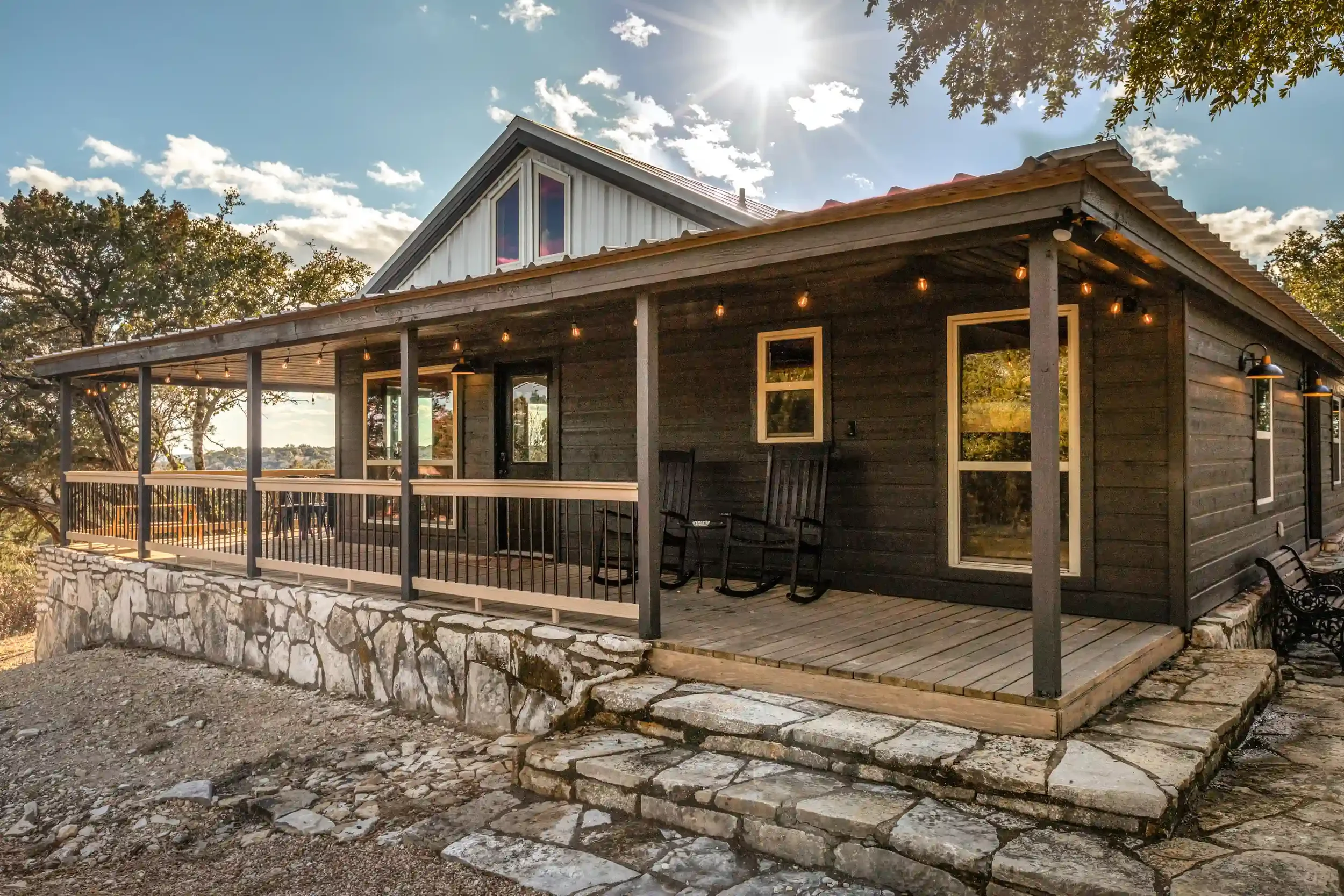 A modern house with a wooden exterior and a sloped roof, featuring large windows, a spacious back porch with string lights, and a stone foundation. There are trees and a blue sky with some clouds in the background.