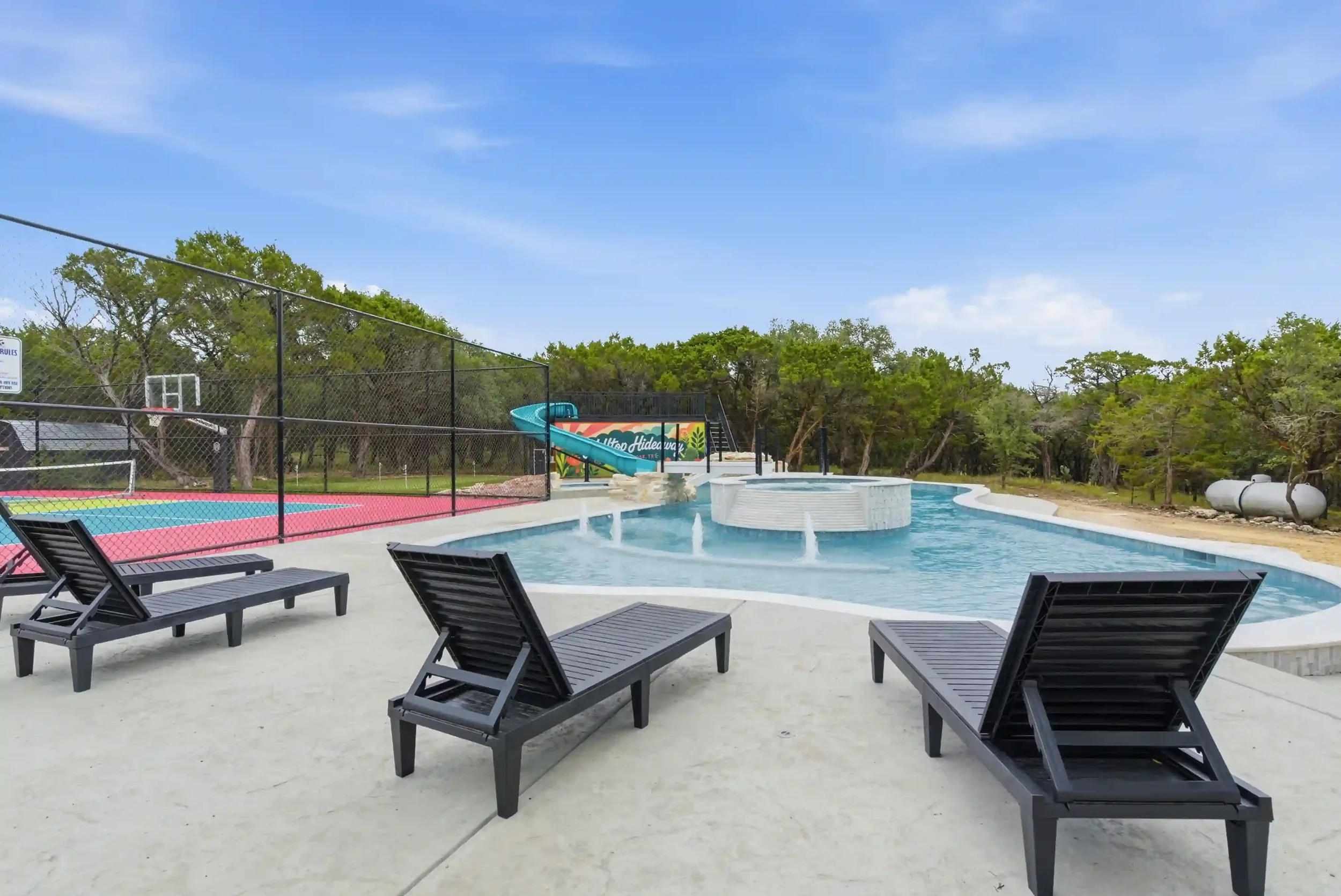 A swimming pool with lounge chairs around it, a waterslide, and trees in the background on a clear, sunny day.