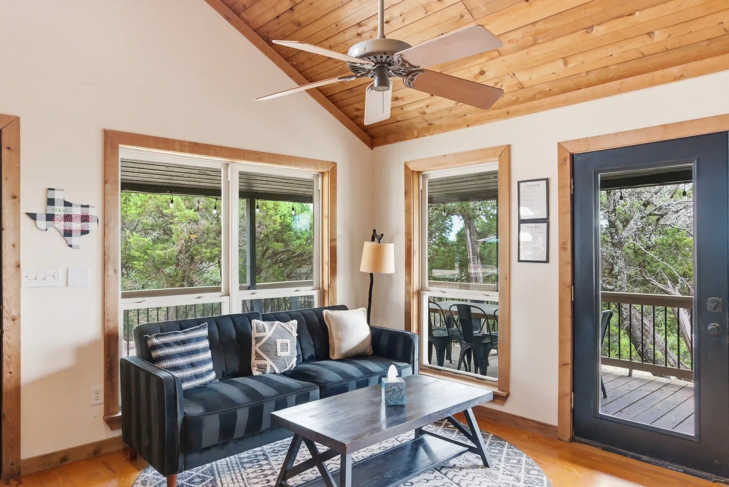 A living room with large windows showing trees outside. The ceiling and window frames are made of wood. There is a dark blue sofa with striped and patterned pillows, a light-colored floor lamp, a rectangular coffee table with a tissue box, and a deco
