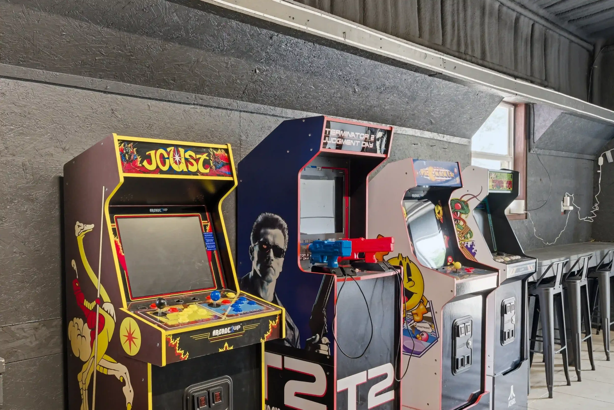 A row of vintage arcade video game machines along a grey textured wall and a window, with power cords plugged into electrical outlets.
