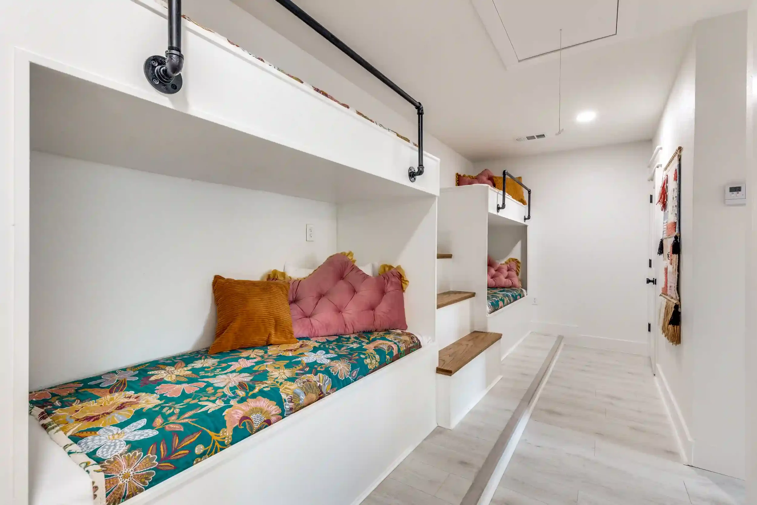 Bunk beds with floral bedding, pink pillows, and wooden stairs in a minimalist white room with a small window and wall decorations.