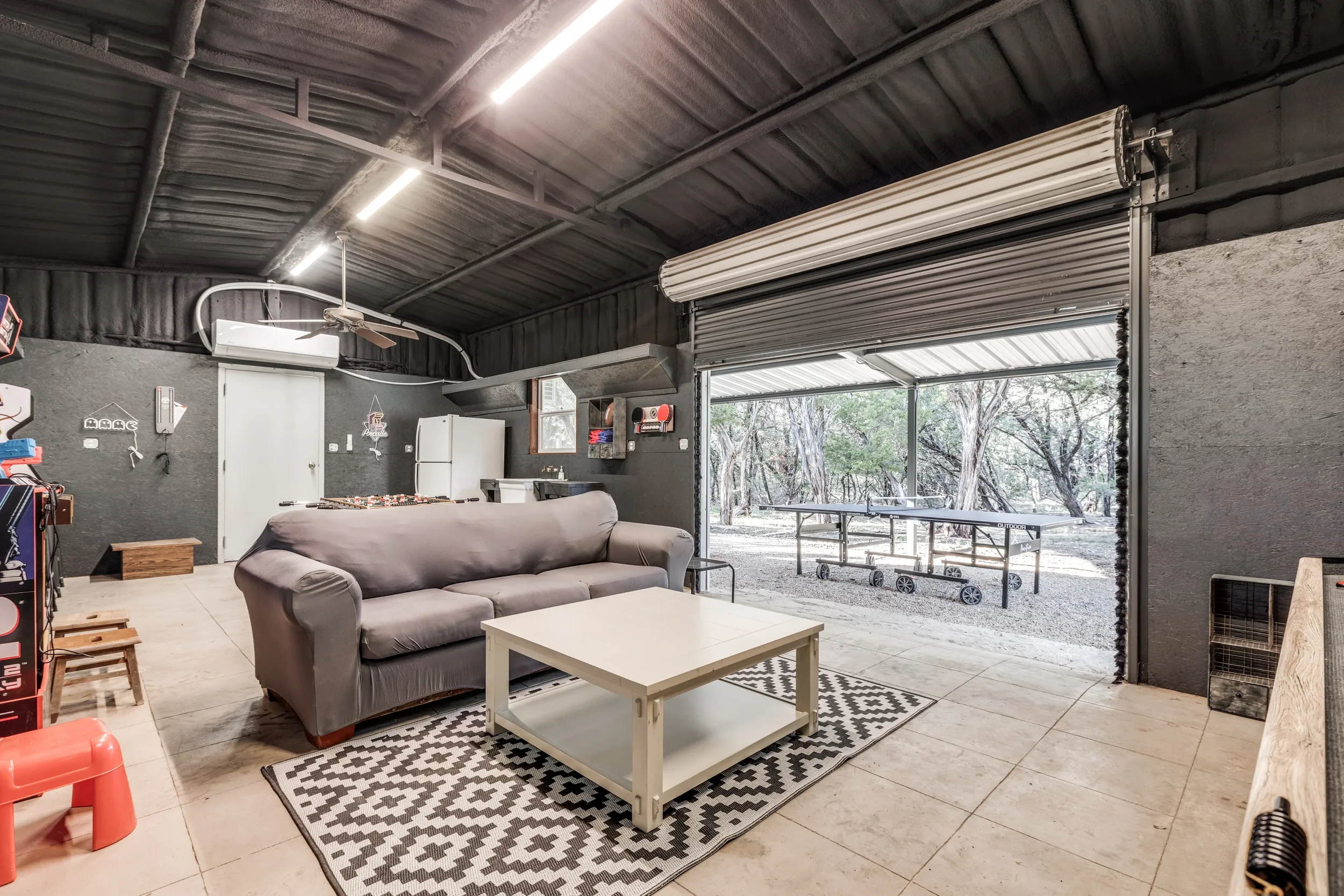 A garage with a gray couch, a white coffee table, and a patterned rug. The garage has an open roll-up door with a view of trees and outdoor furniture. There is a ceiling fan, a white refrigerator, and various items on the walls and shelves.