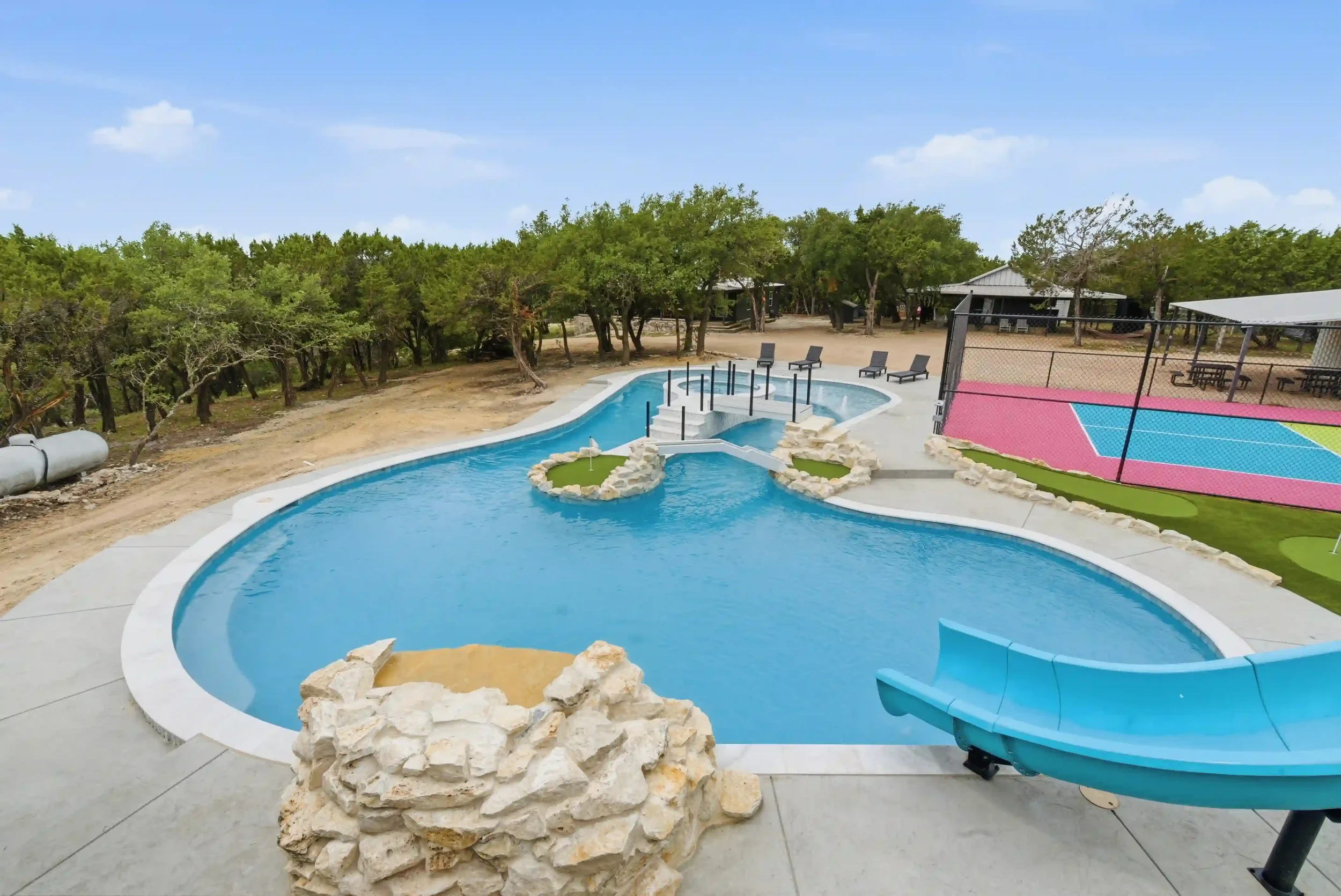 Empty swimming pool with a waterslide, surrounded by trees, in an outdoor recreational area with a basketball court and picnic tables.