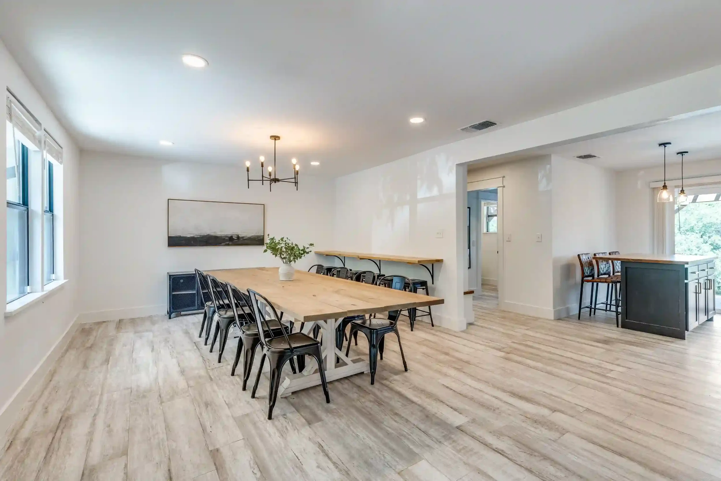 Interior view of a modern dining room with a long wooden table, black chairs, white walls, large windows, pendant lighting, and some artwork on the walls.