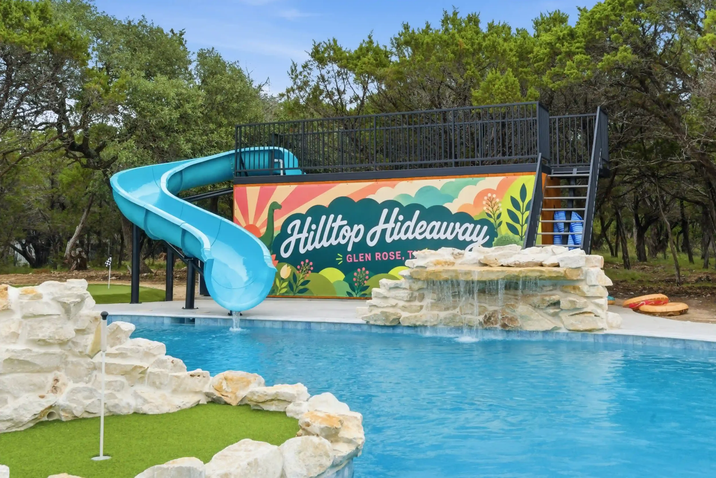 A water slide at Hilltop Hideaway in Glen Rose, Texas, with a painted mural, a pool with decorative rocks, and surrounded by lush green trees.