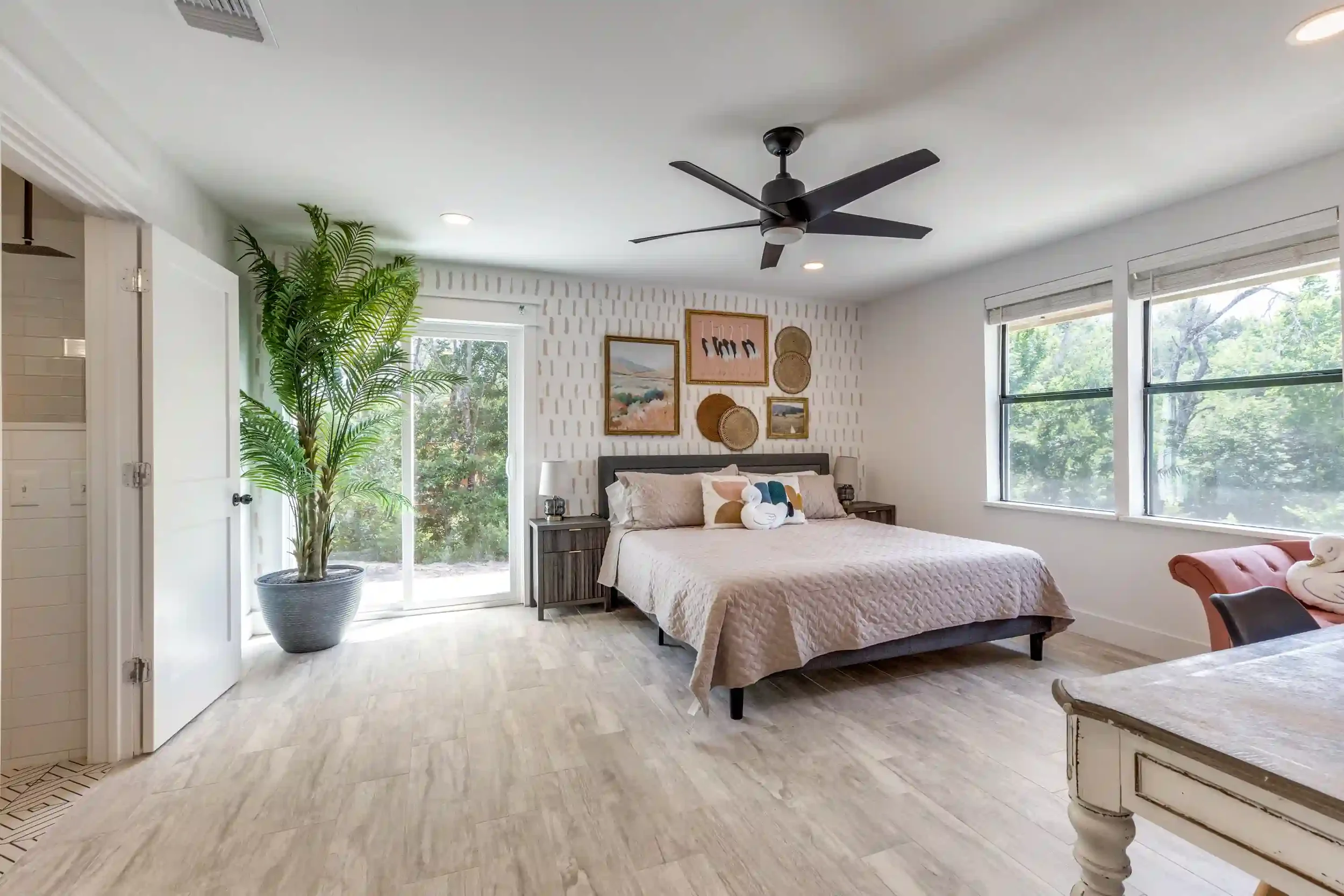 A bedroom with a large bed, framed wall art, and windows showing trees outside. There is a tall green plant near a sliding door, and the room has ceiling lights and a black ceiling fan.