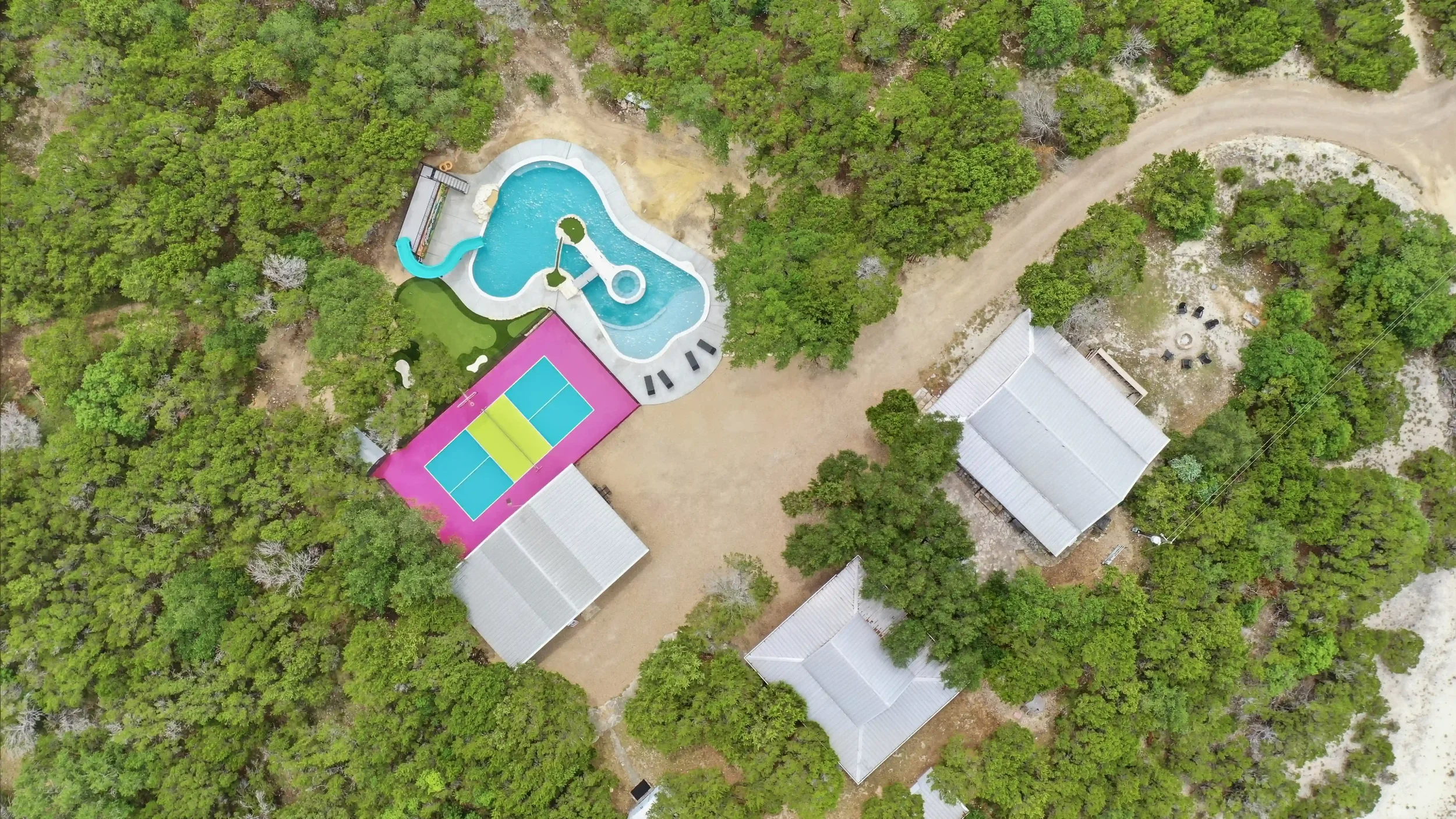 An aerial view of a backyard with a resort-style pool featuring a water slide, surrounded by trees and multiple buildings, including a tennis court and residences.