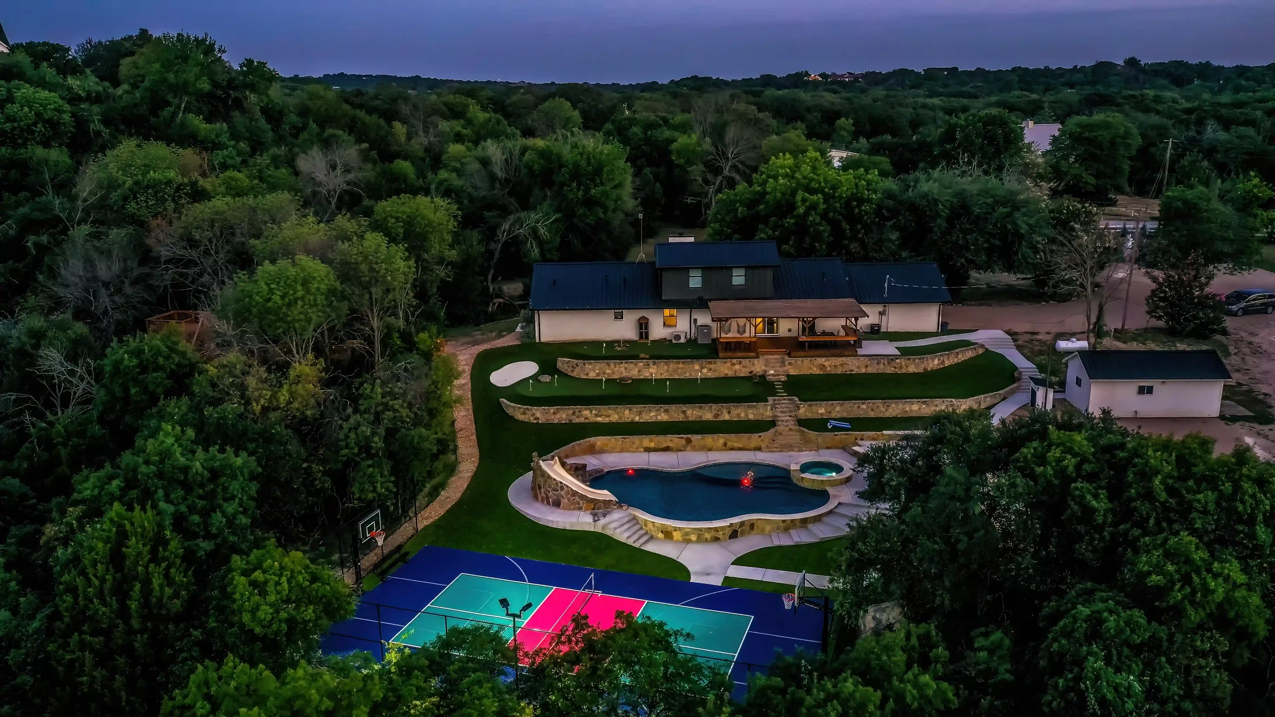 Aerial view of a backyard with a swimming pool, multiple trees, a basketball court, a house with a deck, and a sports court, surrounded by lush greenery and wooded area.