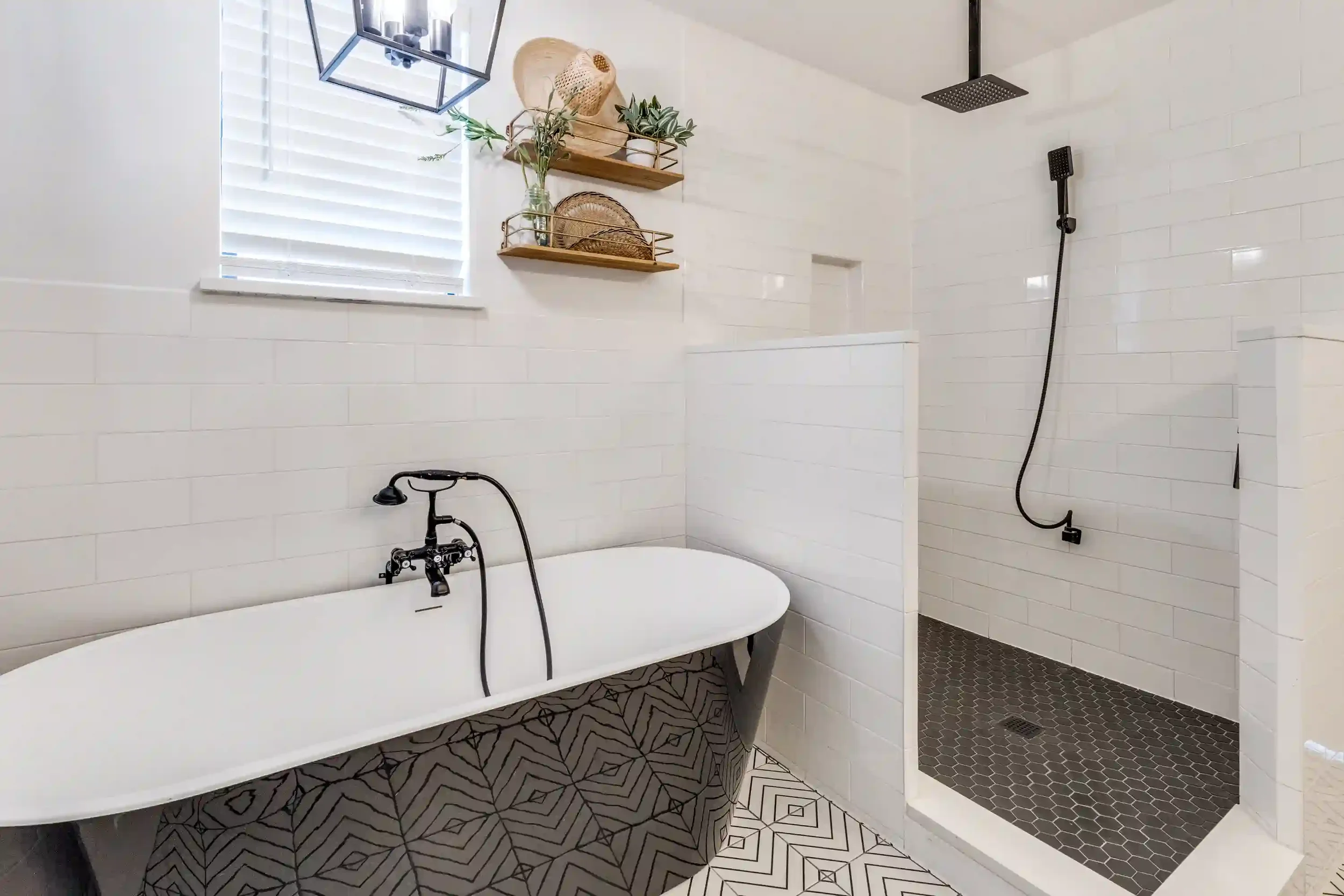 Modern bathroom with white tiled walls, a freestanding bathtub, a black showerhead, a window with white blinds, wooden shelves with plants and baskets, and patterned floor tiles.