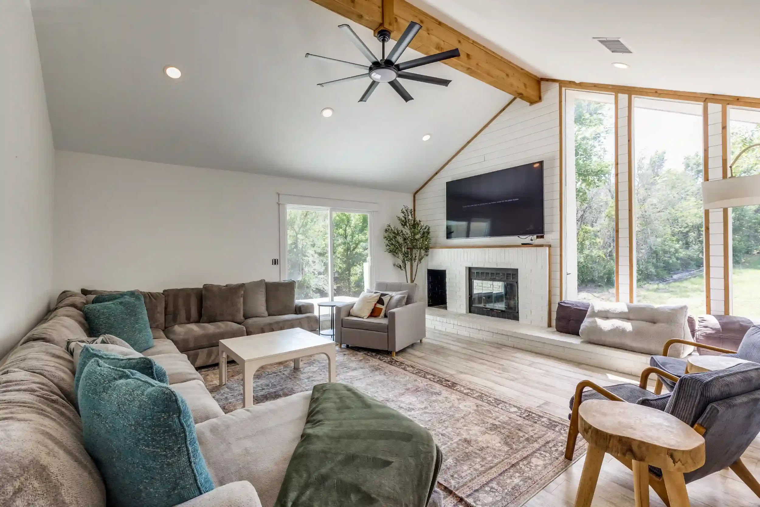 Living room with a large beige sectional sofa with various teal and beige pillows, a small white coffee table, a gray armchair, a fireplace, a wall-mounted TV, large windows showing trees outside, wood-paneled ceiling, and a ceiling fan.