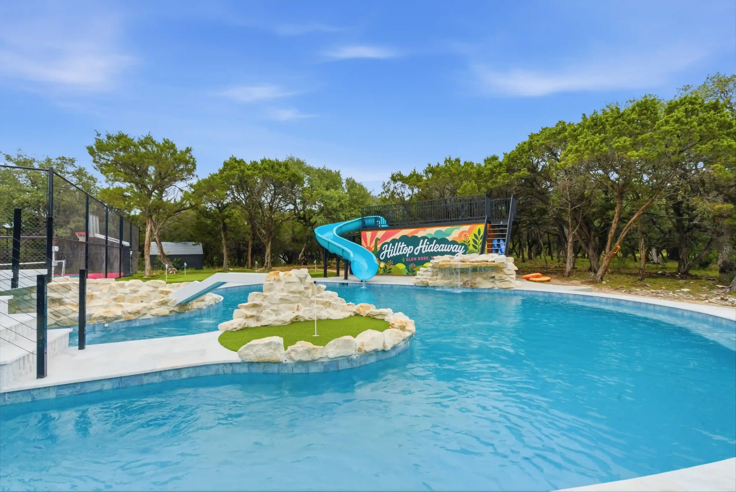 A water park with a blue water slide and a small pool. There are artificial rocks and a sign that reads "Hilltop Hideaway" in the background, surrounded by trees and a clear blue sky.