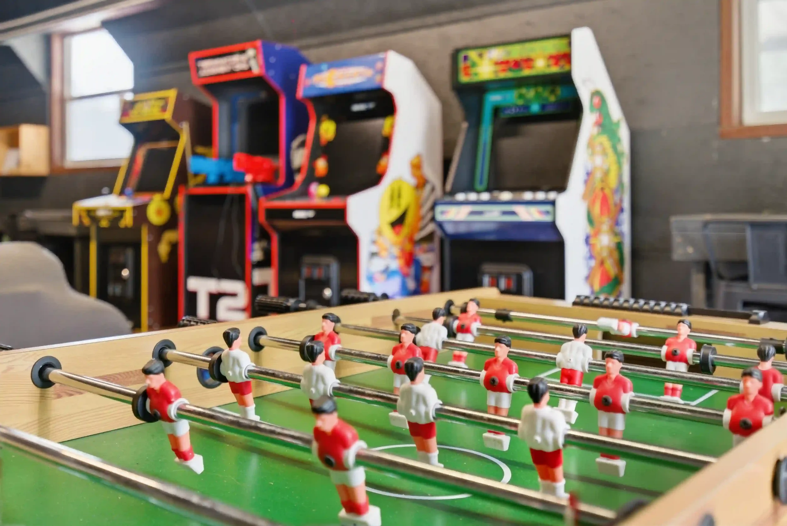 A close-up of a foosball table with small plastic players in red and white uniforms, set on green grass-like surface, with vintage arcade game machines in the background against a gray wall.