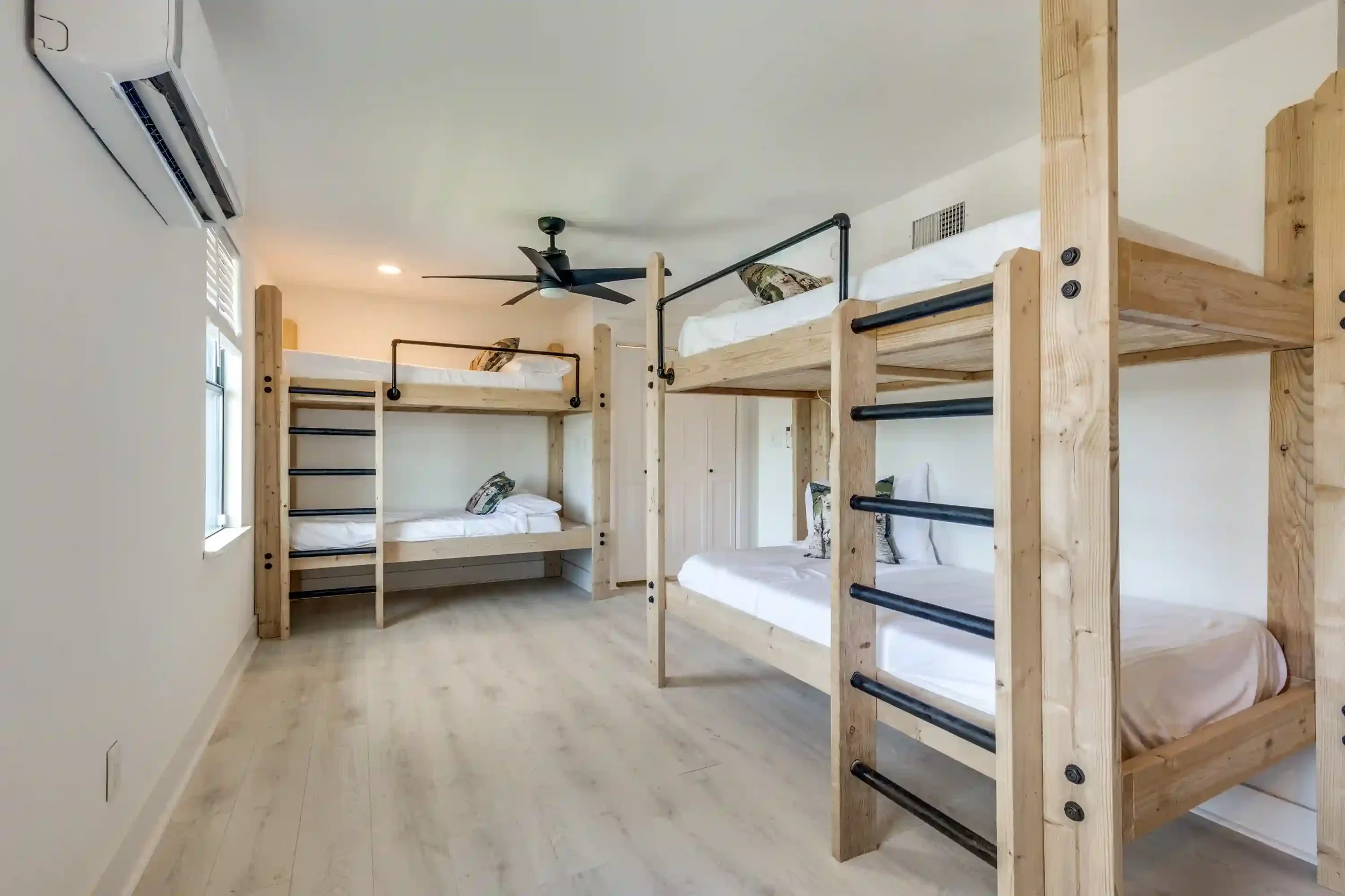 Indoor room with four wooden bunk beds with black metal ladders, white bedding, and pillows. Ceiling fan, air conditioning unit on left wall, window letting in natural light, light wood flooring, cream walls.