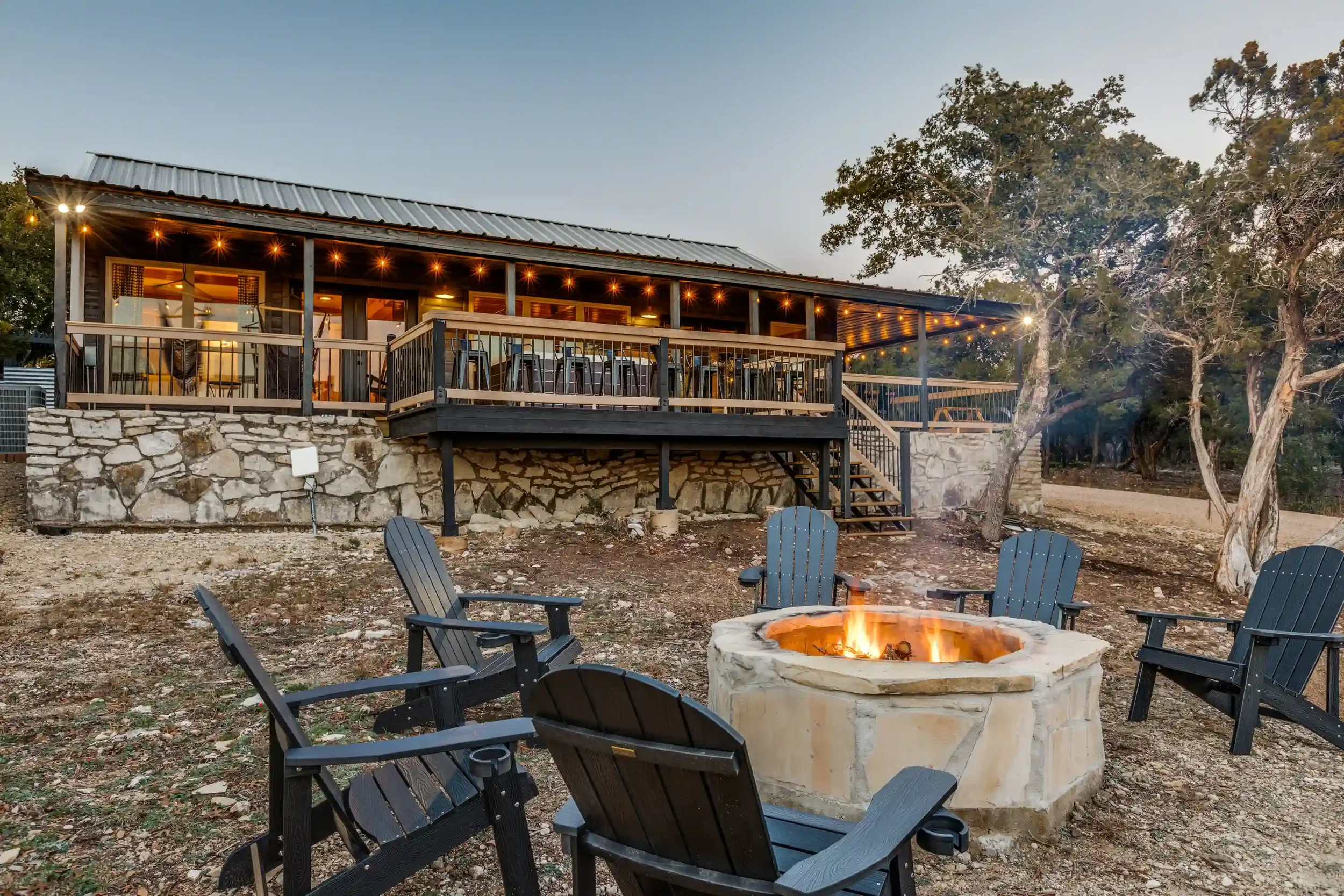 A rustic outdoor deck with string lights, outdoor chairs around a fire pit, and a house with a balcony surrounded by trees at sunset.