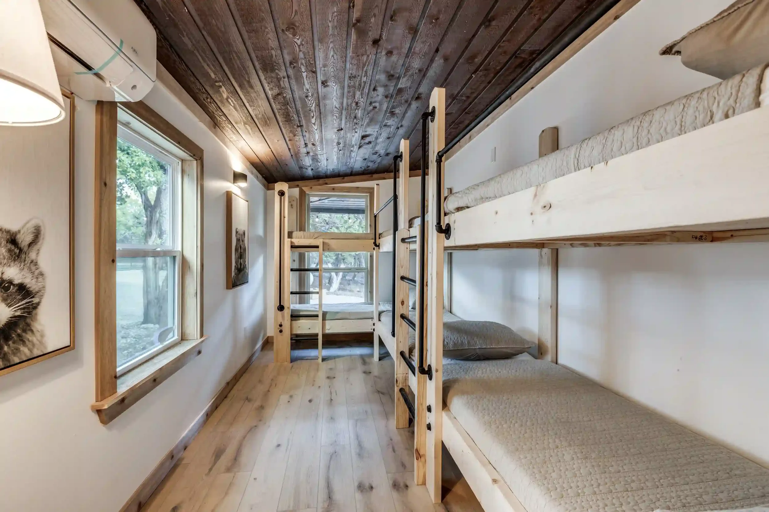 A cozy room with wooden flooring and walls, featuring built-in bunk beds with beige mattresses and black metal ladders, large windows allowing natural light, and a dark wood vaulted ceiling.