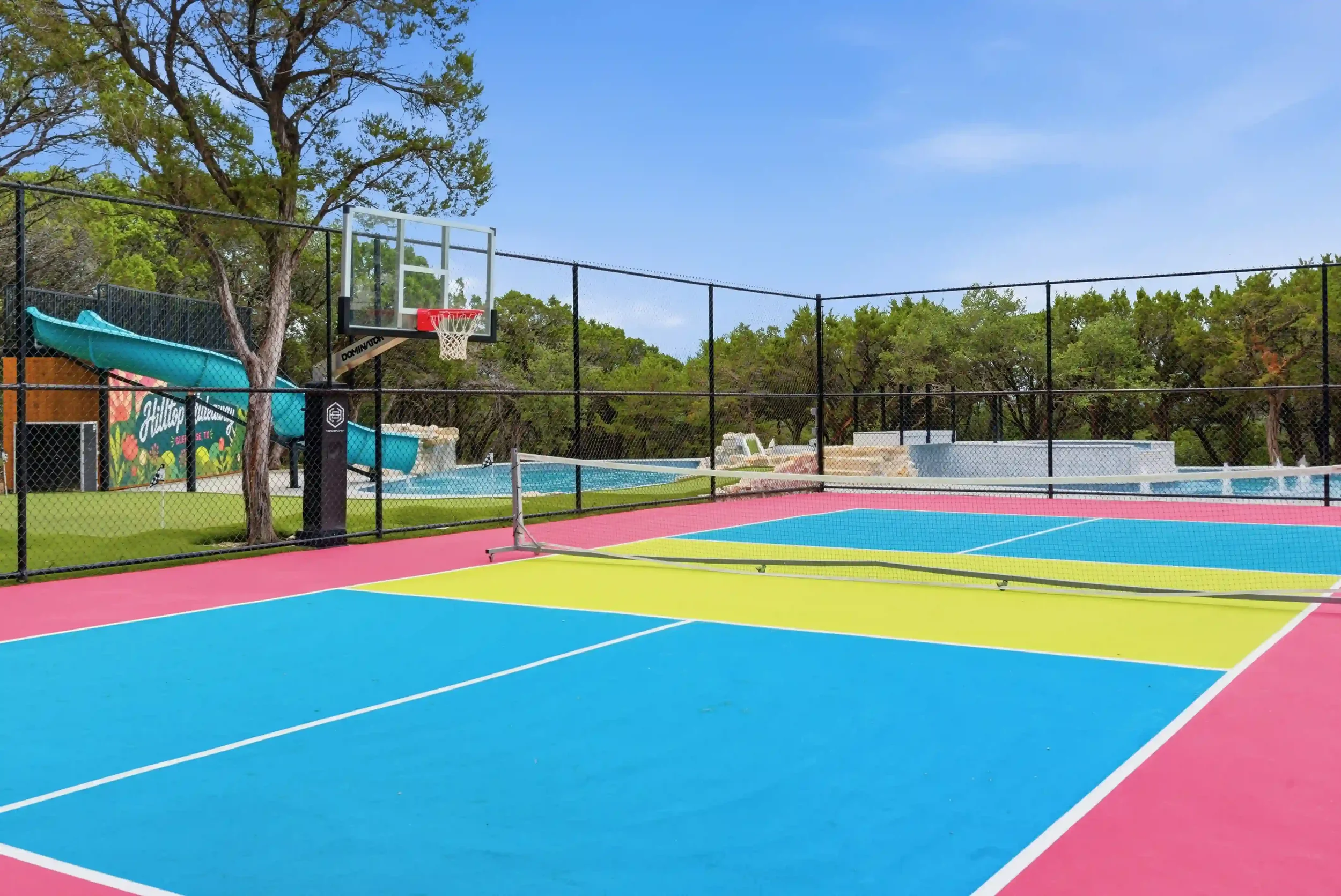 Colorful outdoor basketball and tennis court with a basketball hoop, surrounded by green trees and a blue sky.