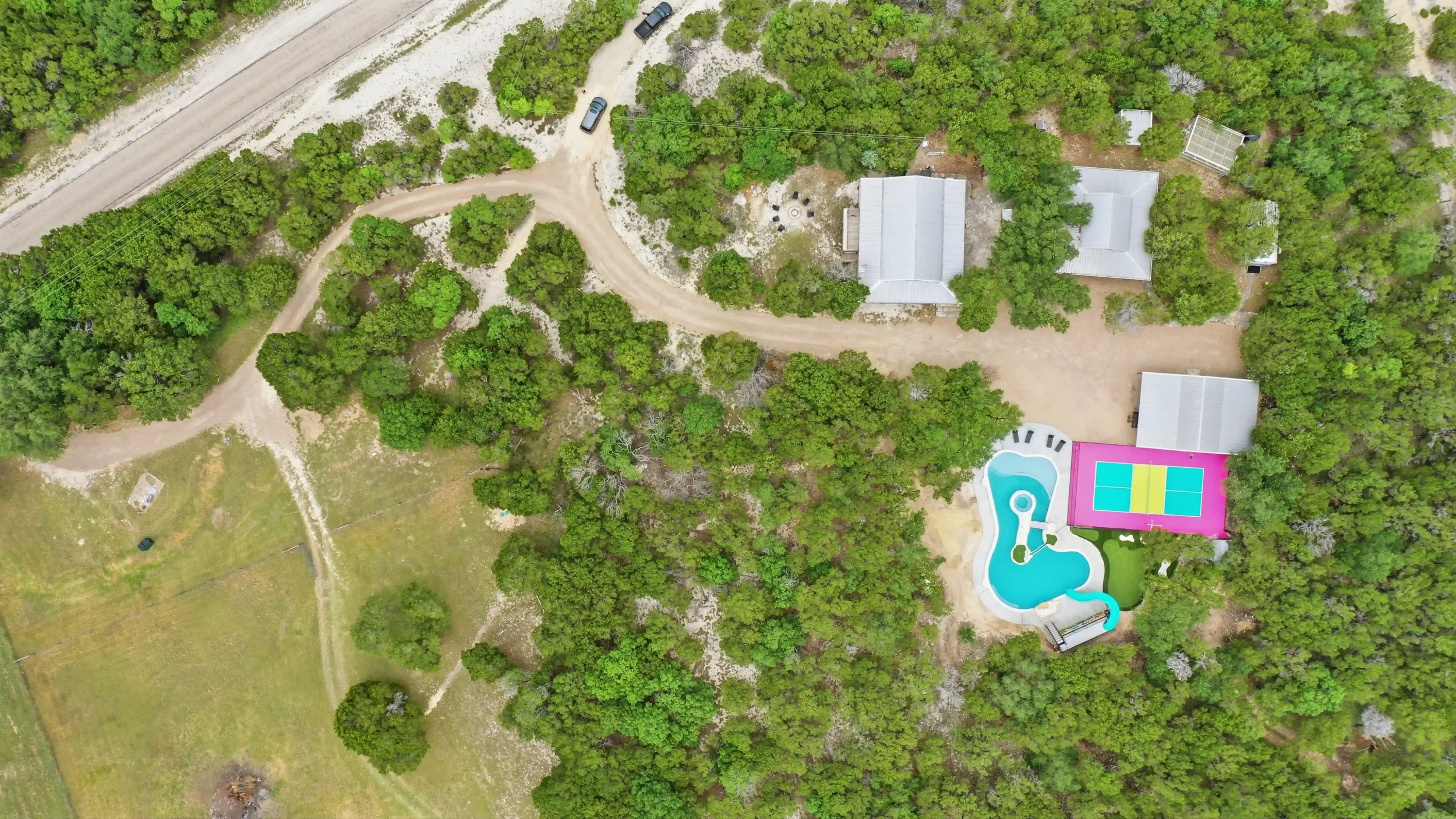 Aerial view of a backyard with a forested area, a house with a gray roof, a colorful pool with a waterslide, and a tennis court.