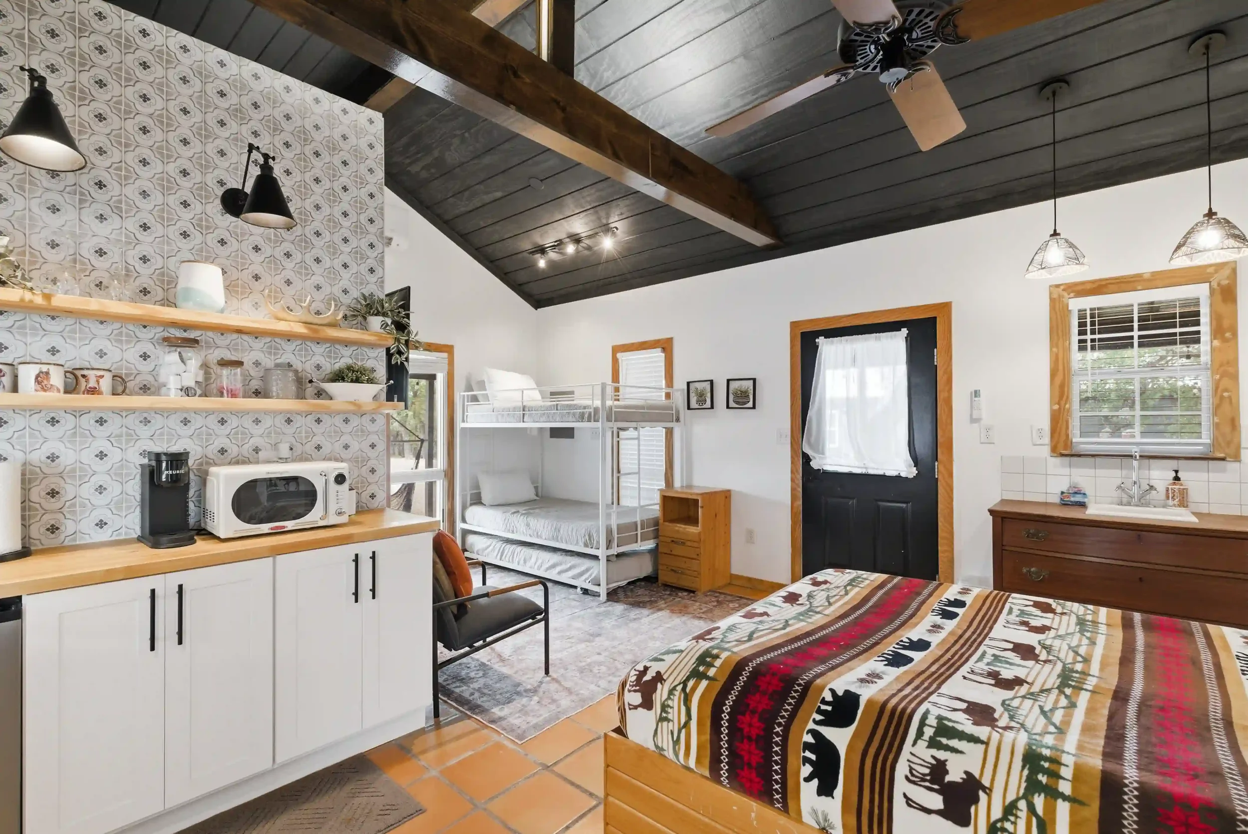 Inside a room with a vaulted dark wood ceiling, ceiling fan, and modern decor. Contains a bunk bed, a kitchen counter with appliances, framed pictures on the wall, and a window with white curtains.