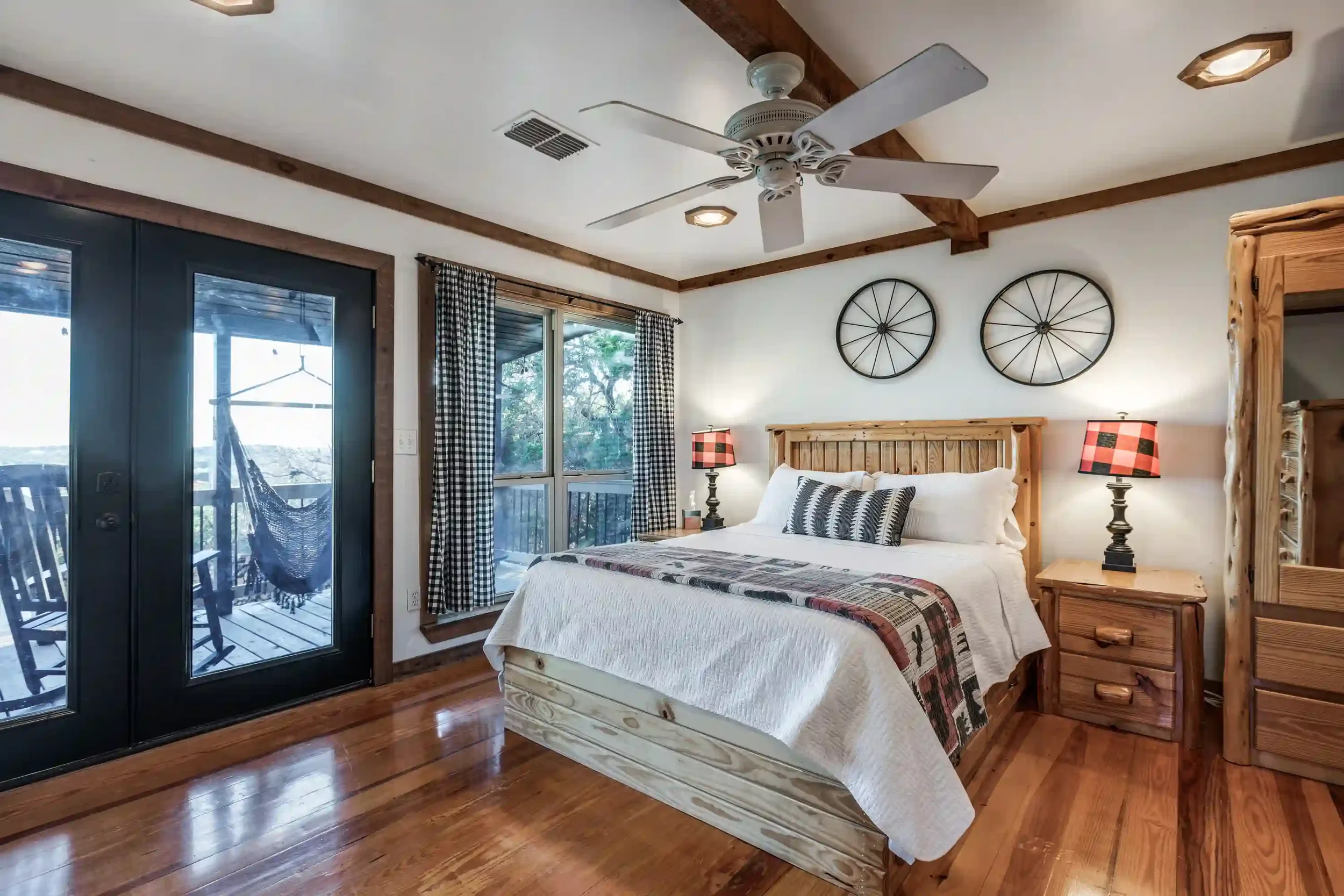 Bedroom with wooden furniture, a white bed with a quilted coverlet and decorative pillows, two matching lamps on bedside tables, a pair of wall decor resembling wheels, and a sliding glass door leading to a balcony with outdoor seating.