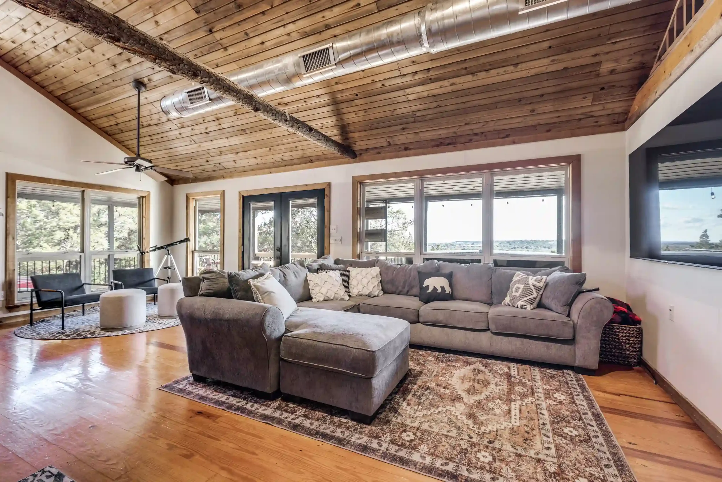 A living room with a wooden ceiling and large windows showing a view outside. The room has a large sectional sofa with pillows, a rug, a telescope near the windows, and a ceiling fan.