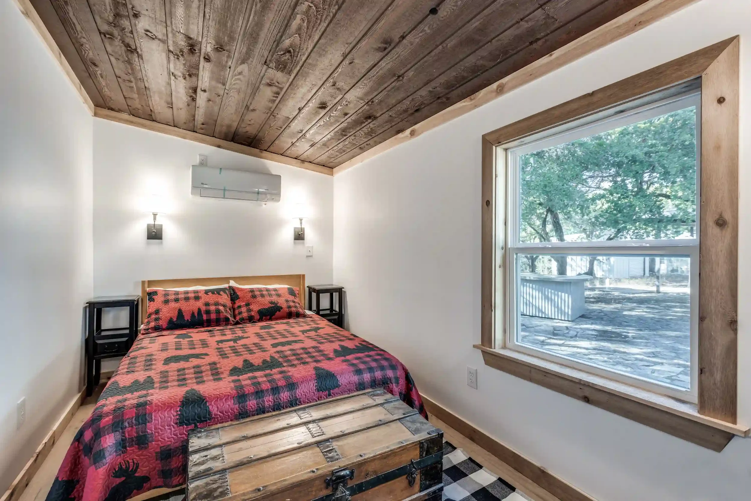 A bedroom with a wooden ceiling, white walls, and a large window showing trees outside. There is a bed with a red and black log cabin quilt, black side tables, and black lamps. An industrial-style wooden chest is at the foot of the bed, and the floor