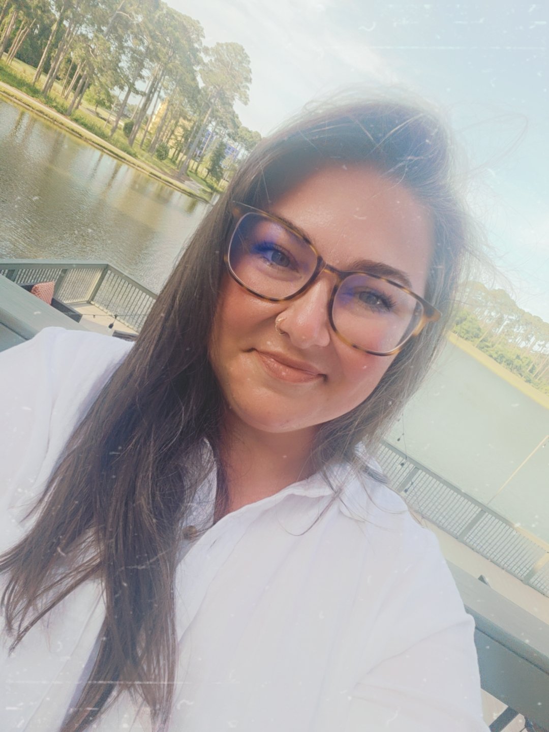 A woman with long brown hair, glasses, and a nose ring taking a selfie outdoors by a body of water with trees and a fence in the background.