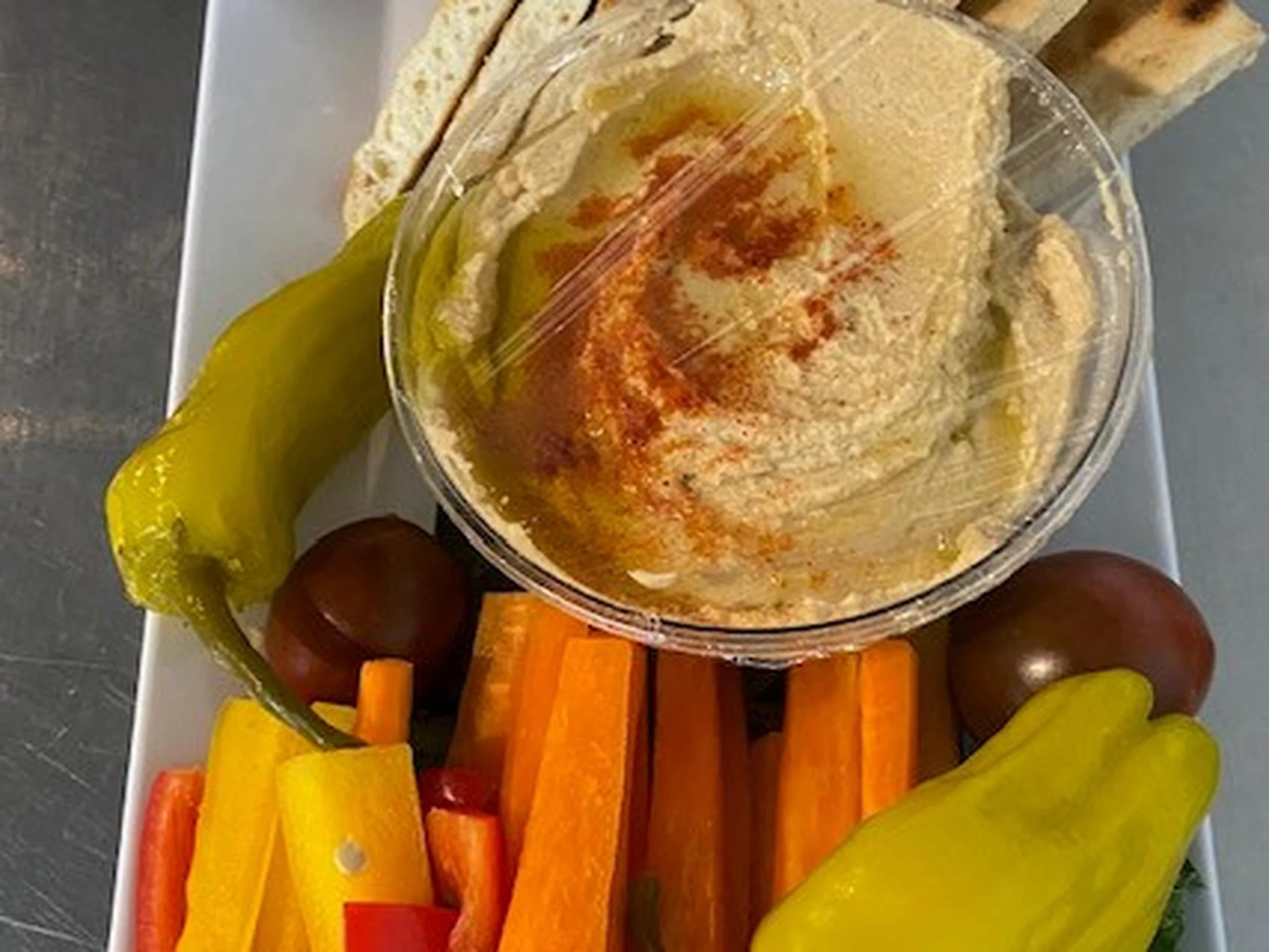 A clear bowl of hummus topped with paprika, olive oil, and spices, surrounded by sliced yellow and red bell peppers, carrots, and green grapes placed on a white rectangular plate.