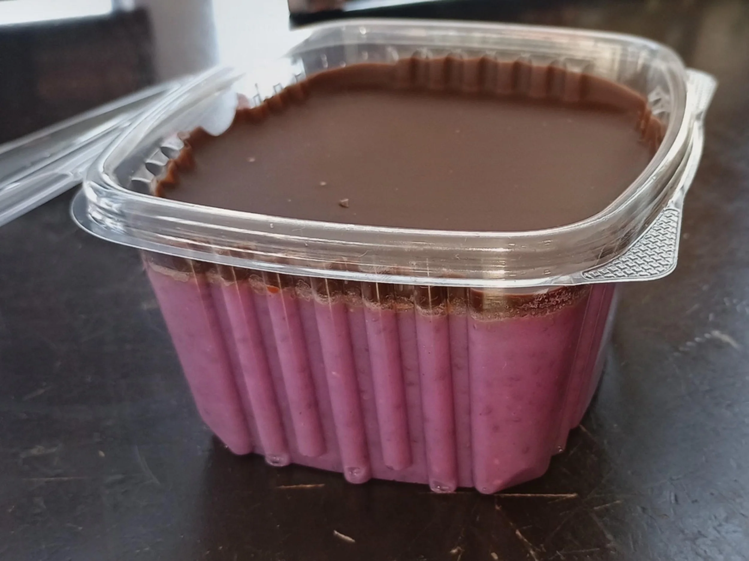 A plastic container filled with a smooth, brown chocolate dessert, with a ridged pink paper liner around the sides.