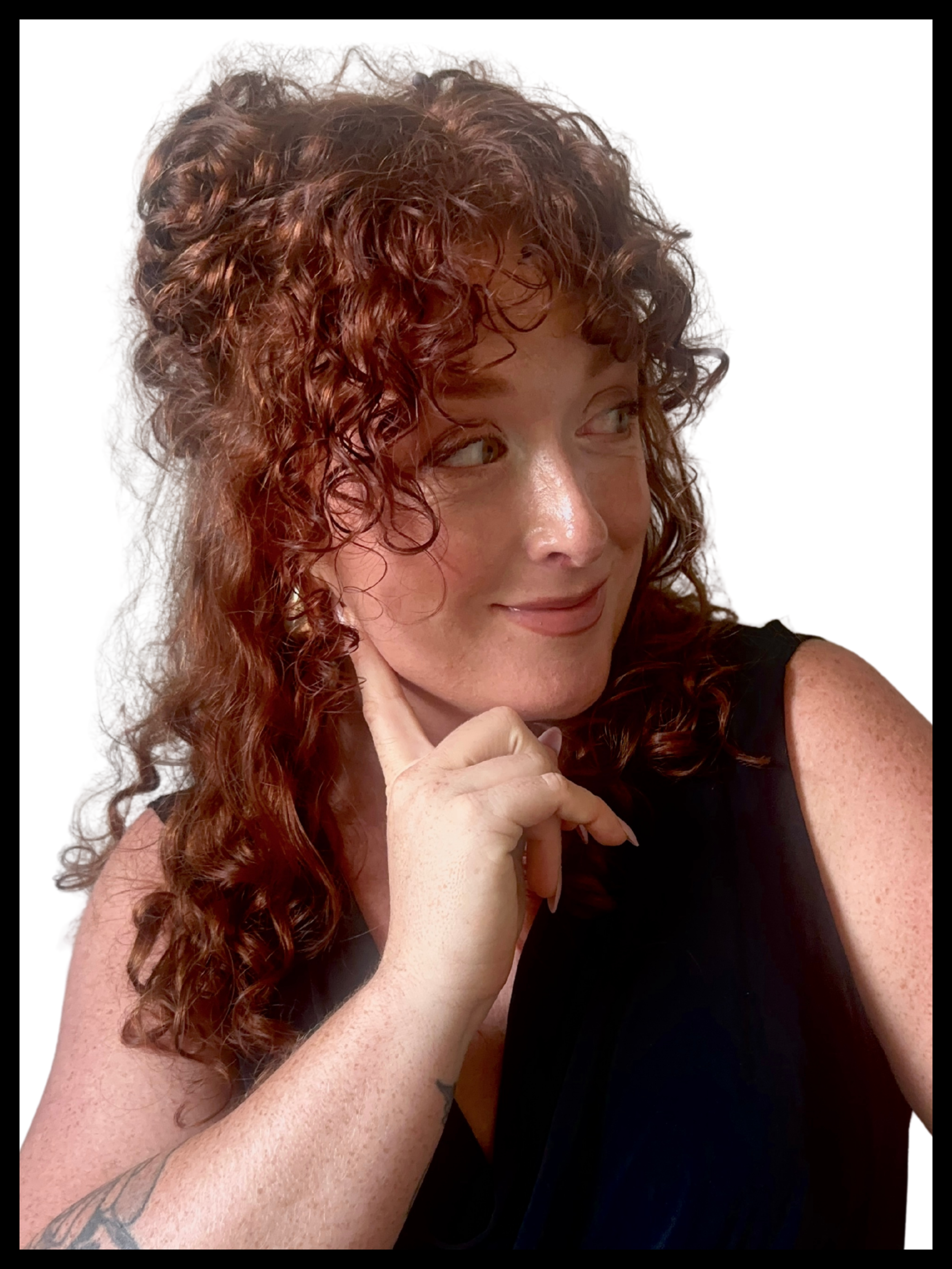 A woman with curly red hair and fair skin is smiling and looking to her left. She has freckles and is wearing a sleeveless black top. She has tattoos on her left arm and a thoughtful expression, with her right hand touching her chin.