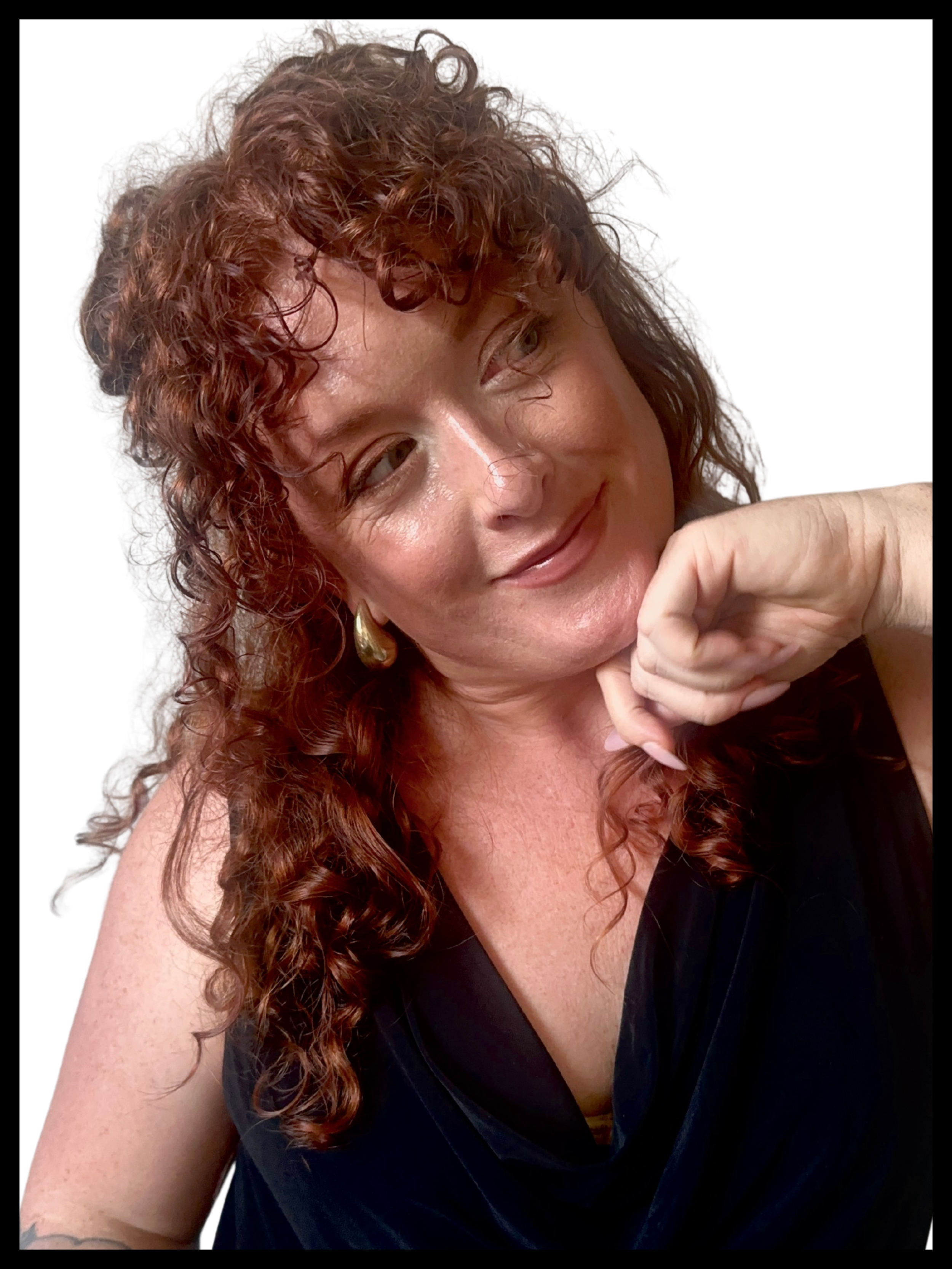 A woman with curly red hair smiling and resting her chin on her hand, wearing a black top and gold earrings, against a white background.