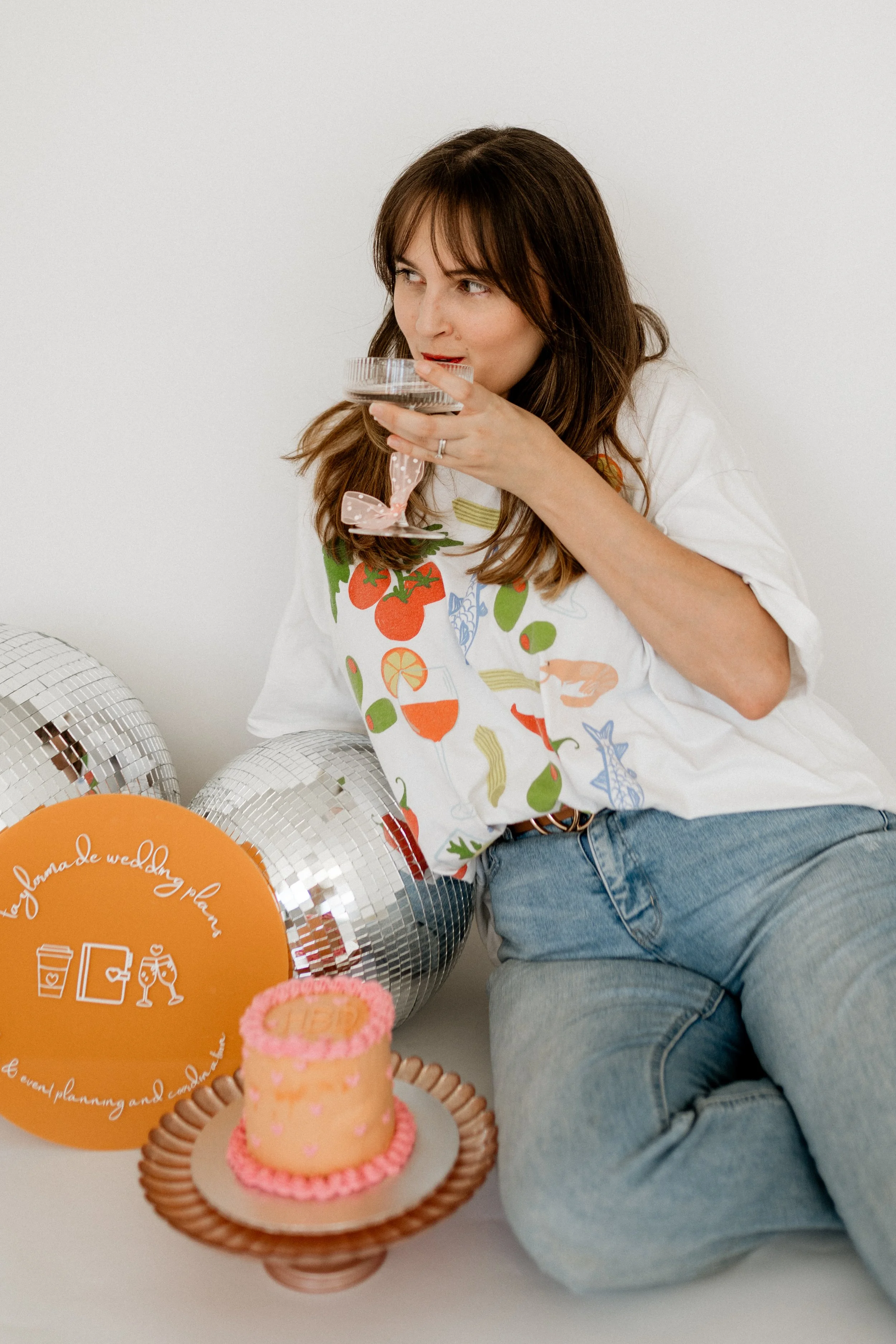 Woman sitting on the floor holding a glass dessert dish, surrounded by balloon decorations and a small cake, celebrating a wedding or event.