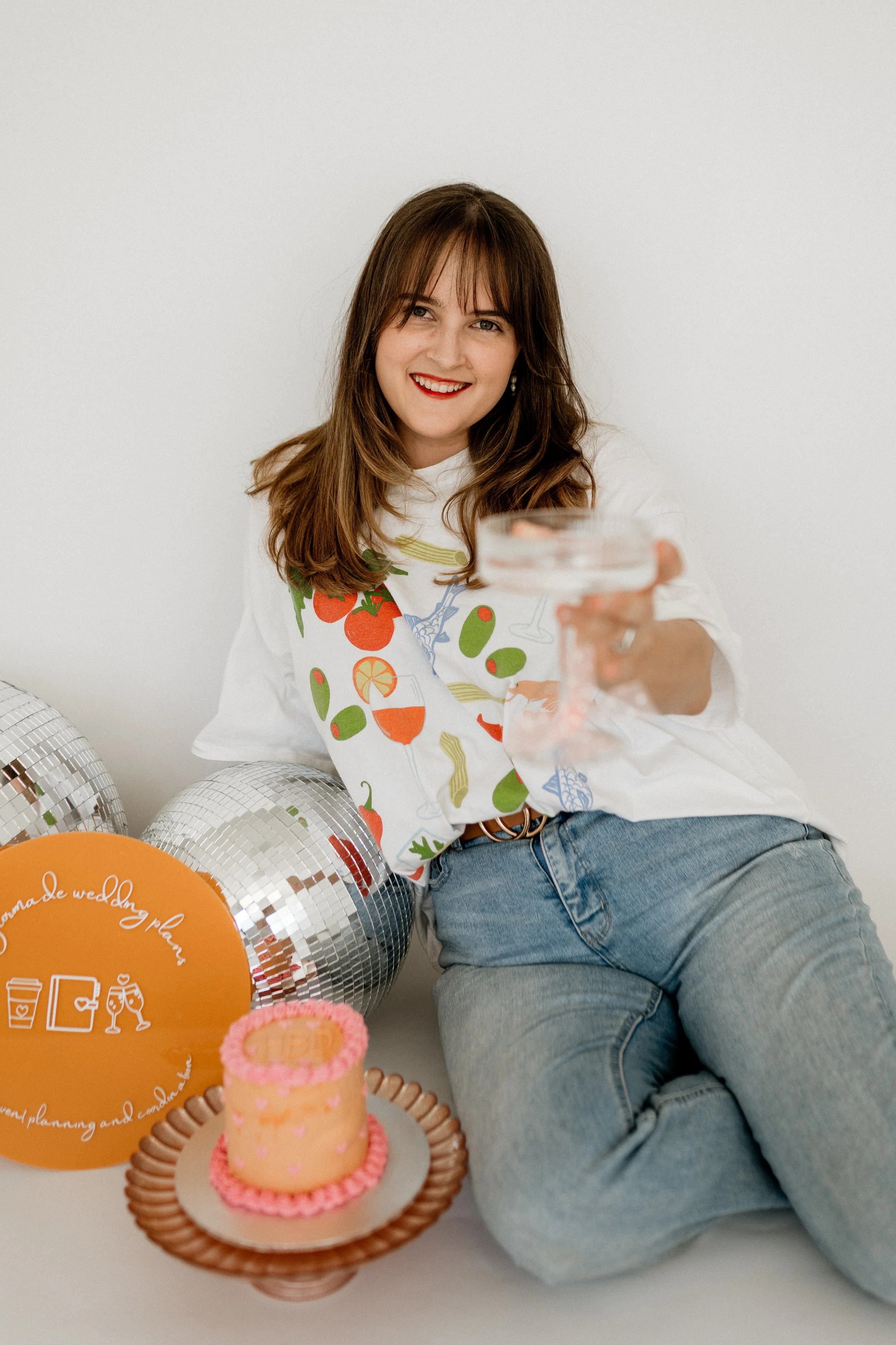 A woman with brown hair and blue eyes, smiling and holding a clear glass, sitting in front of a white wall adorned with helium balloons and decorative items, including a pink and orange birthday cake and a sign with text.