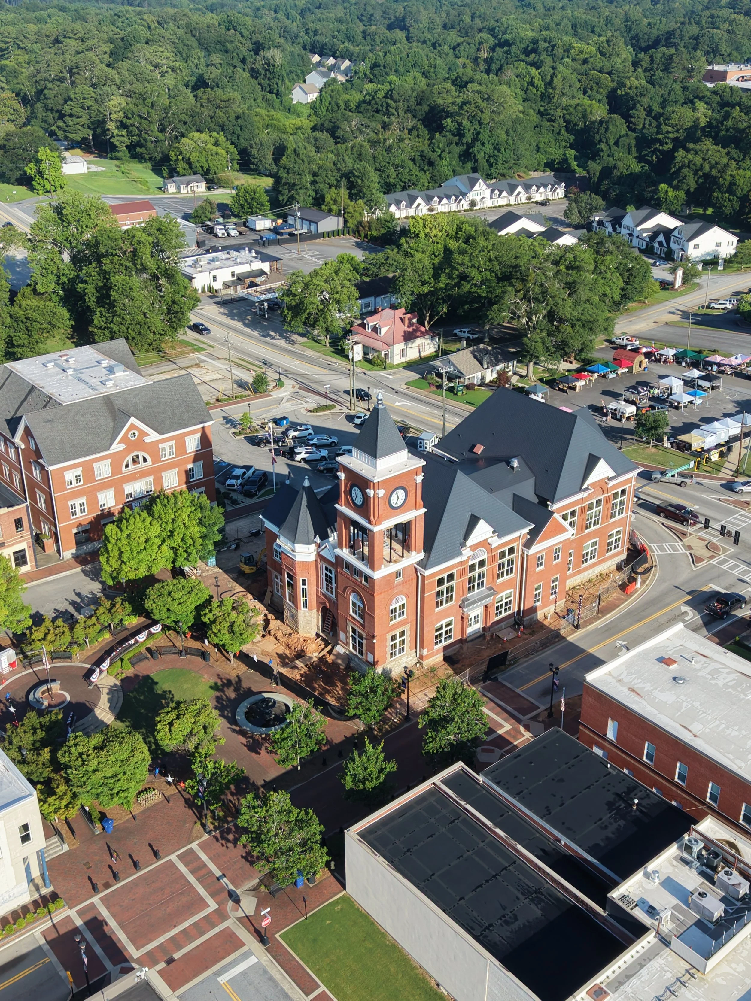 Aerial view of a small town with brick building featuring a clock tower, green trees, small market stalls with tents, parking lots, and surrounding greenery.