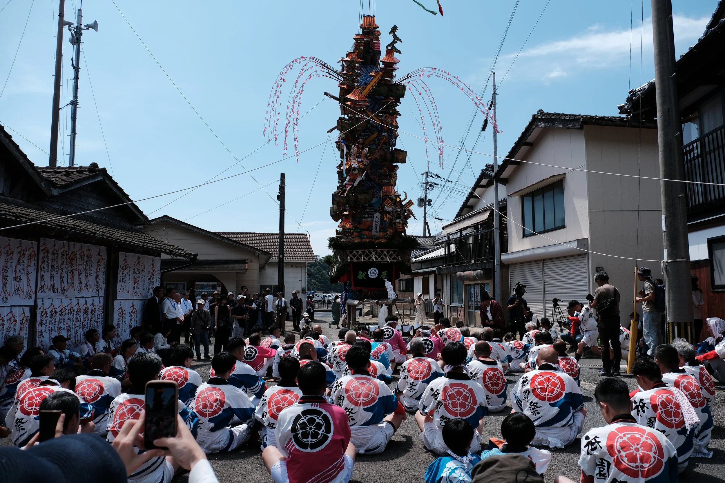 A large, colorful festival float with traditional decorations and architecture, surrounded by a group of people in traditional Japanese clothing, many seated and some standing, with onlookers and photographers capturing the scene in a street.