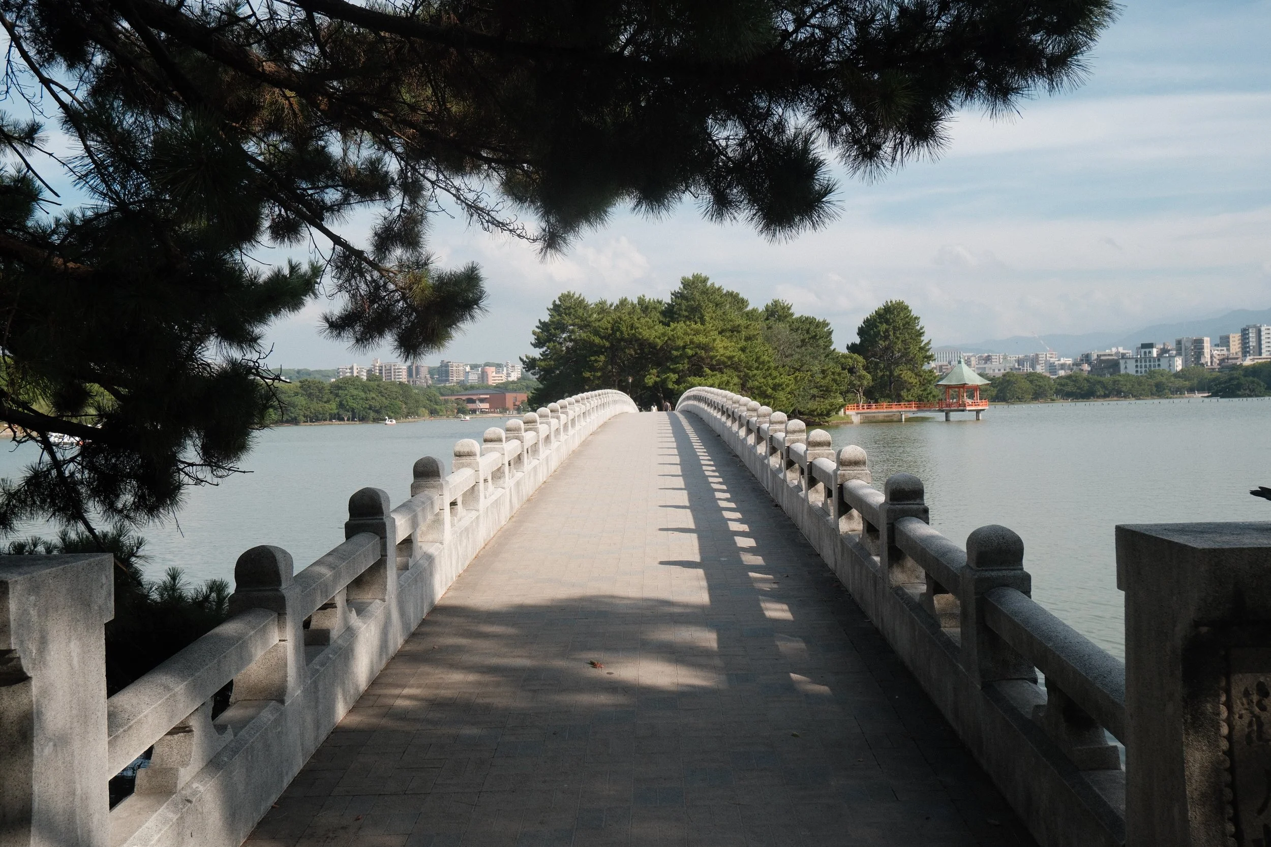 A stone bridge over water leading to a small pavilion with a green roof, surrounded by trees, with city buildings in the distance under a partly cloudy sky. Ohori Park, in Fukuoka City.