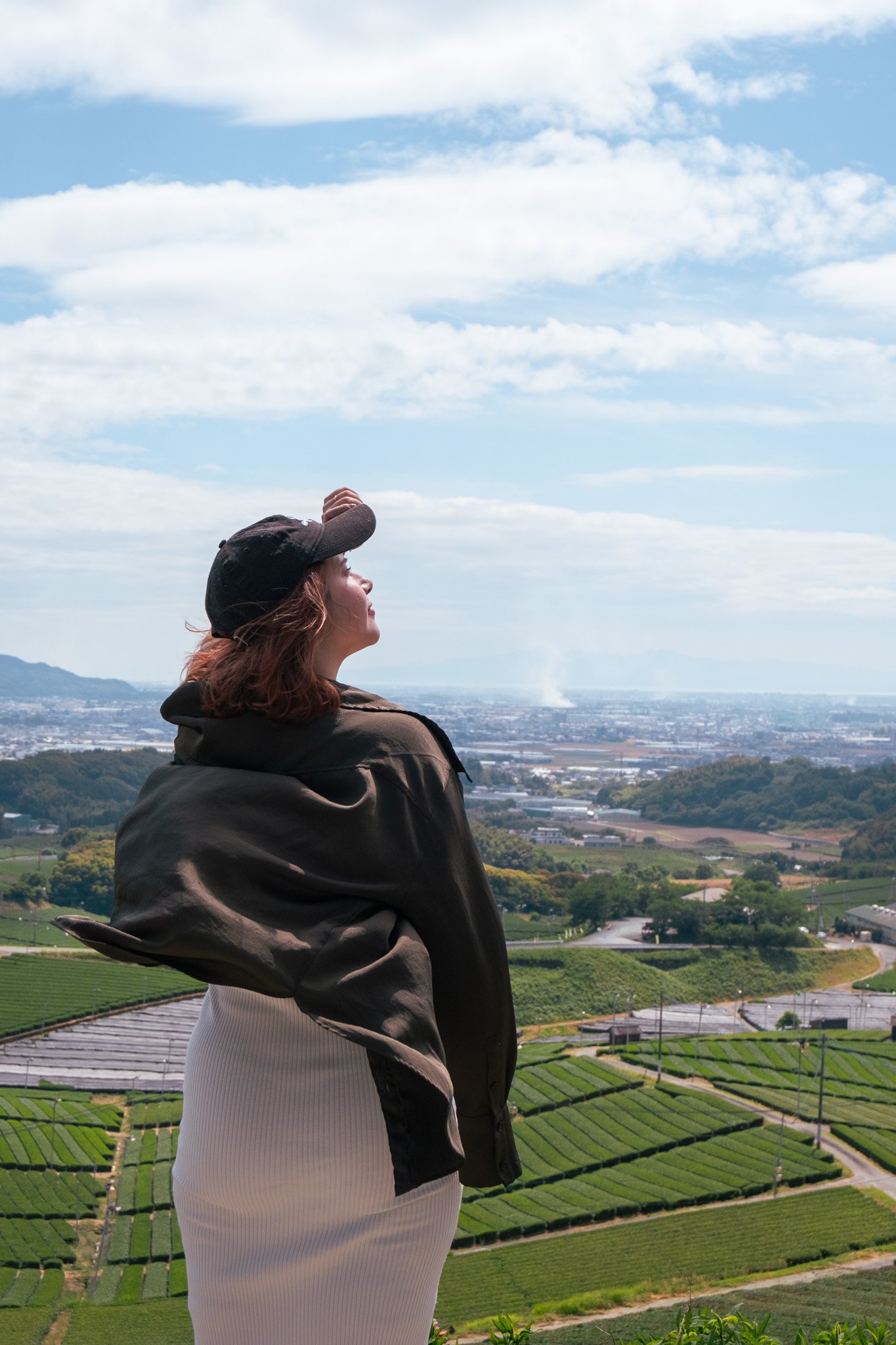 A woman with brown hair wearing a black cap and brown jacket standing outdoors overlooking a landscape of green farmland and a blue sky with white clouds.
