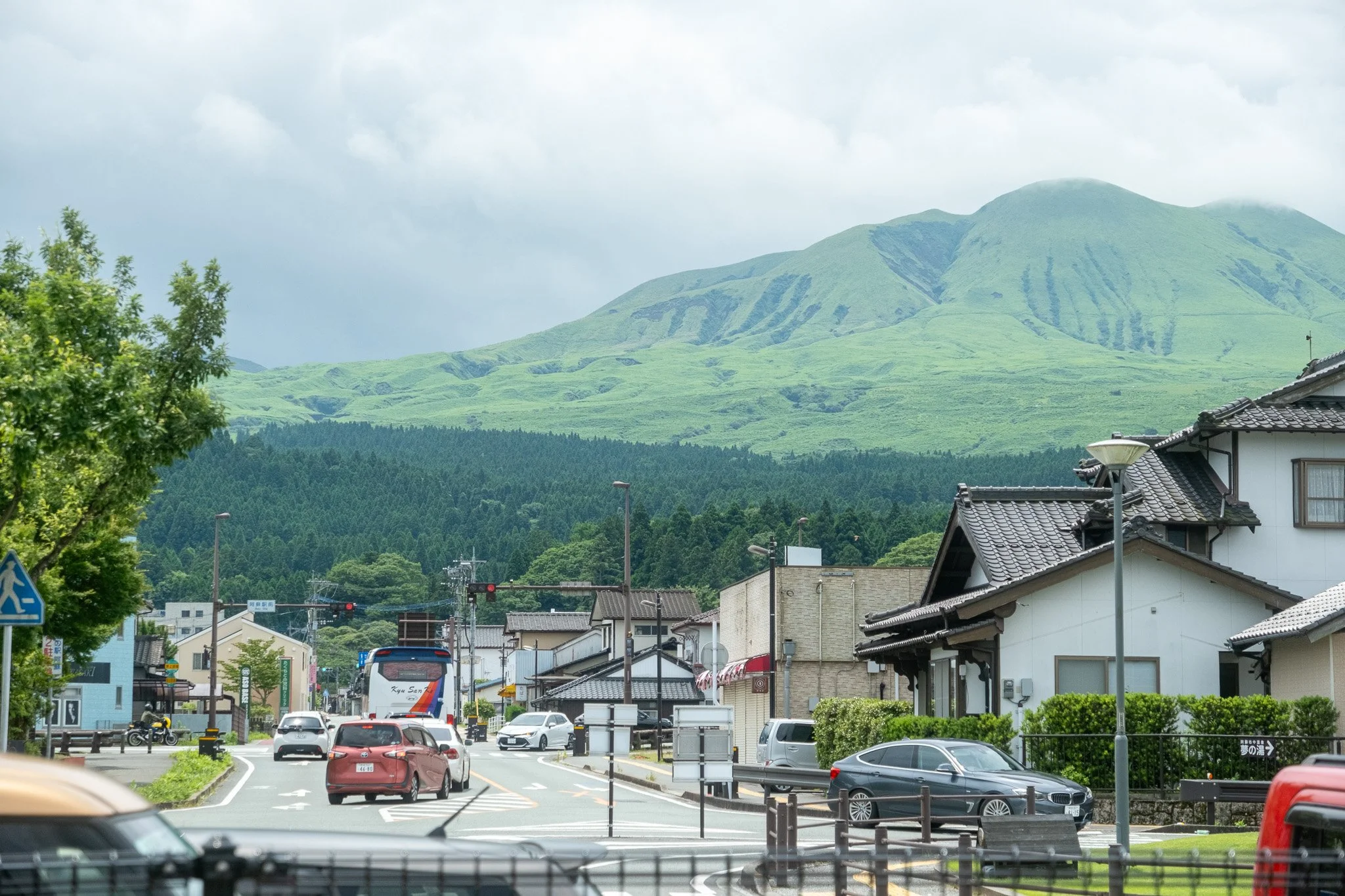 Urban street scene with cars, buildings, and Mt. Aso in the background on a cloudy day.