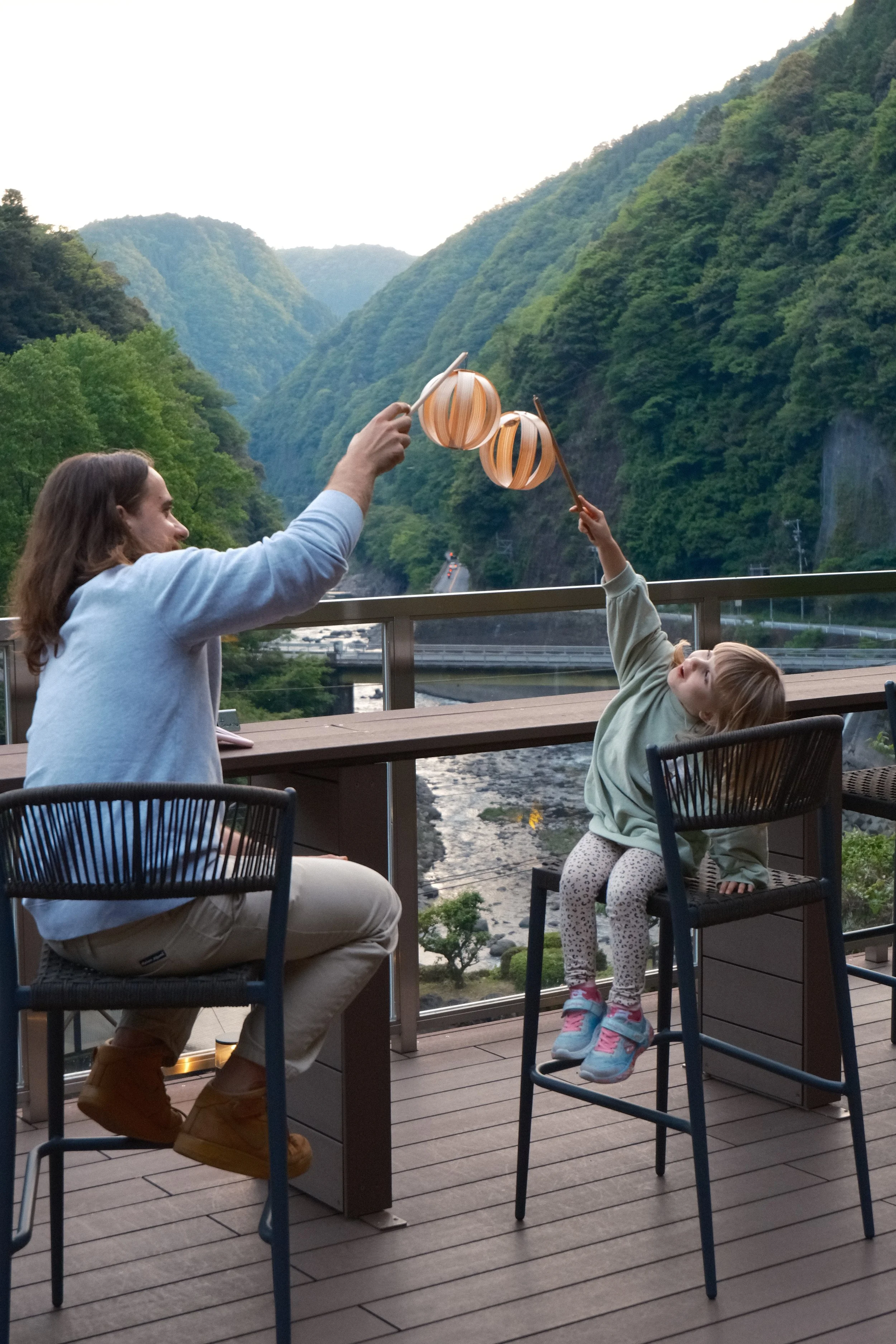 A man and a young girl are outside on a wooden deck overlooking a river in a green mountain valley. They are reaching up to exchange paper lanterns, with the woman holding one lantern and the girl holding the other.