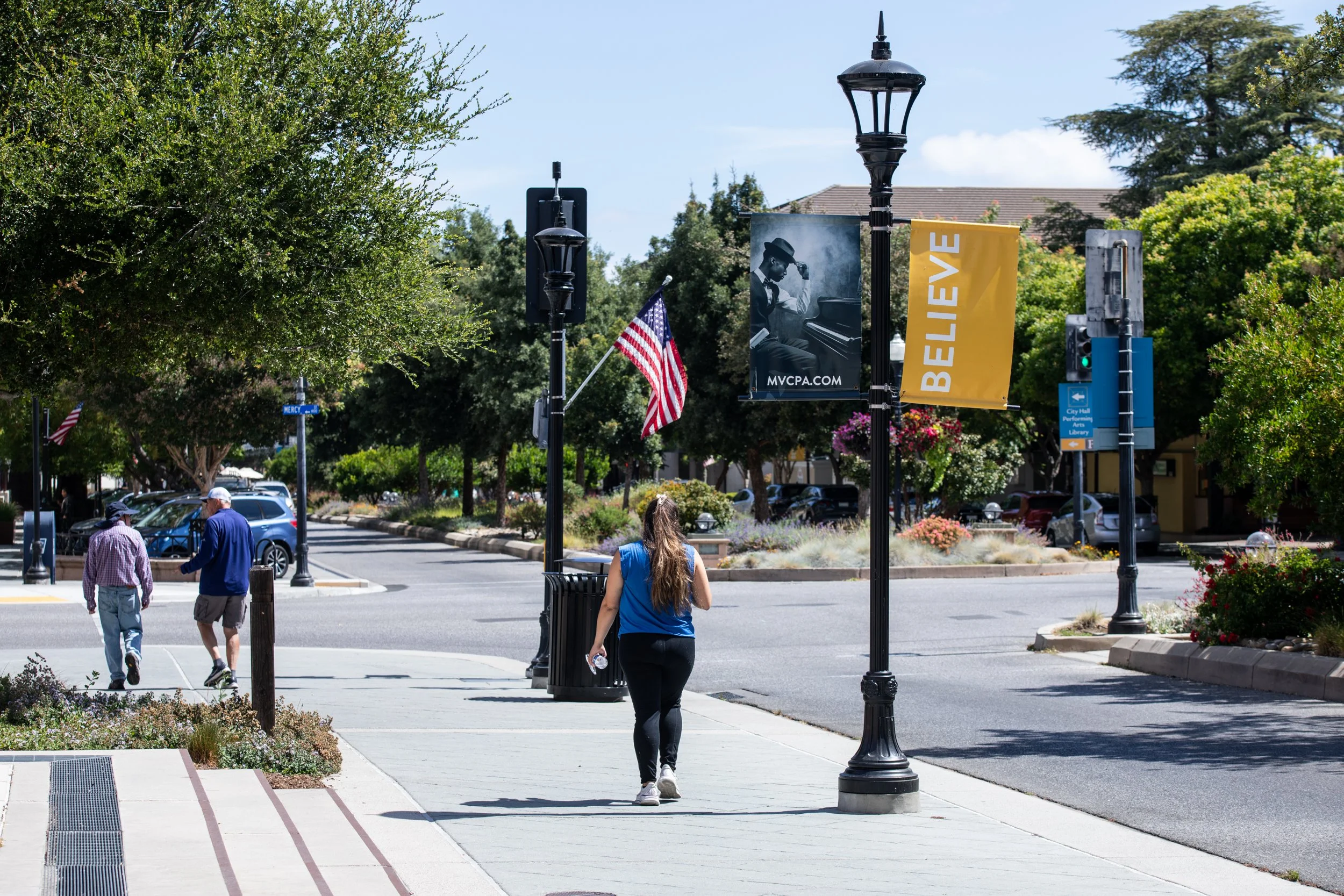 A sidewalk scene with three people walking, street with parked cars, trees, lampposts, banners reading "BELIEVE," American flags, and a building in the background on a sunny day.