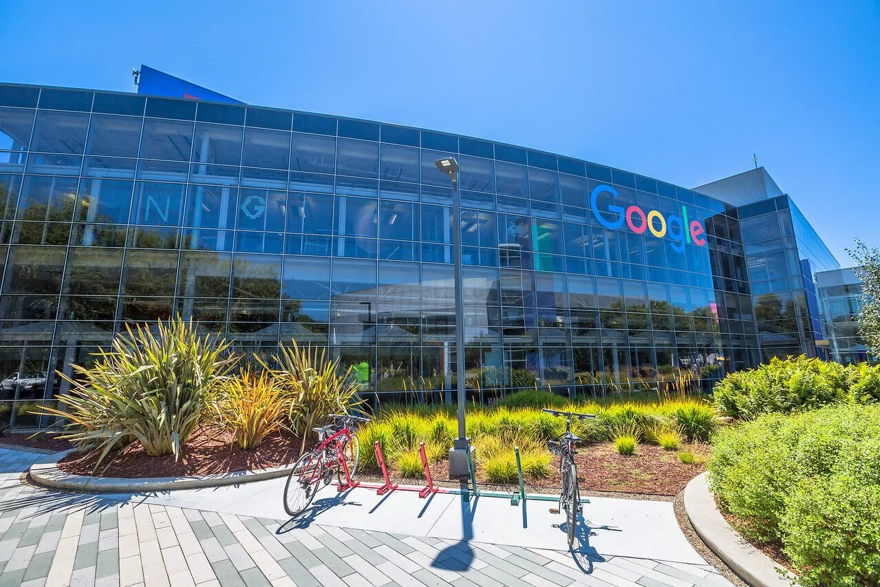 Google headquarters building with a glass exterior, bicycle rack with bikes, landscaped area, and clear blue sky.