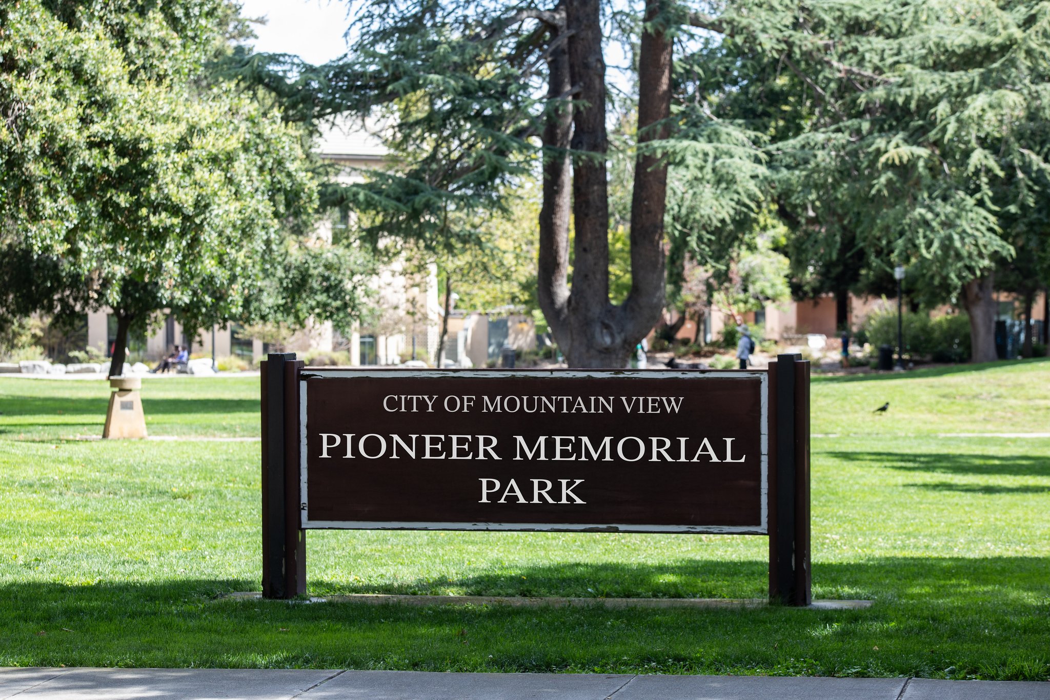 Brown park sign reading 'City of Mountain View Pioneer Memorial Park' in a green park with trees and people in the background.