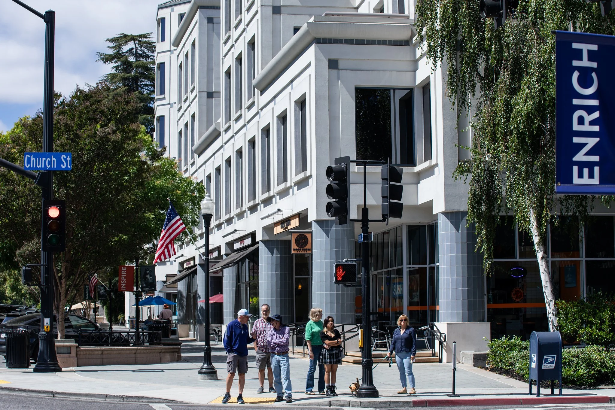 People waiting at a crosswalk on a city street corner in front of a modern white building with large windows. There are street signs, traffic lights, and American flags, with some trees and outdoor seating visible.