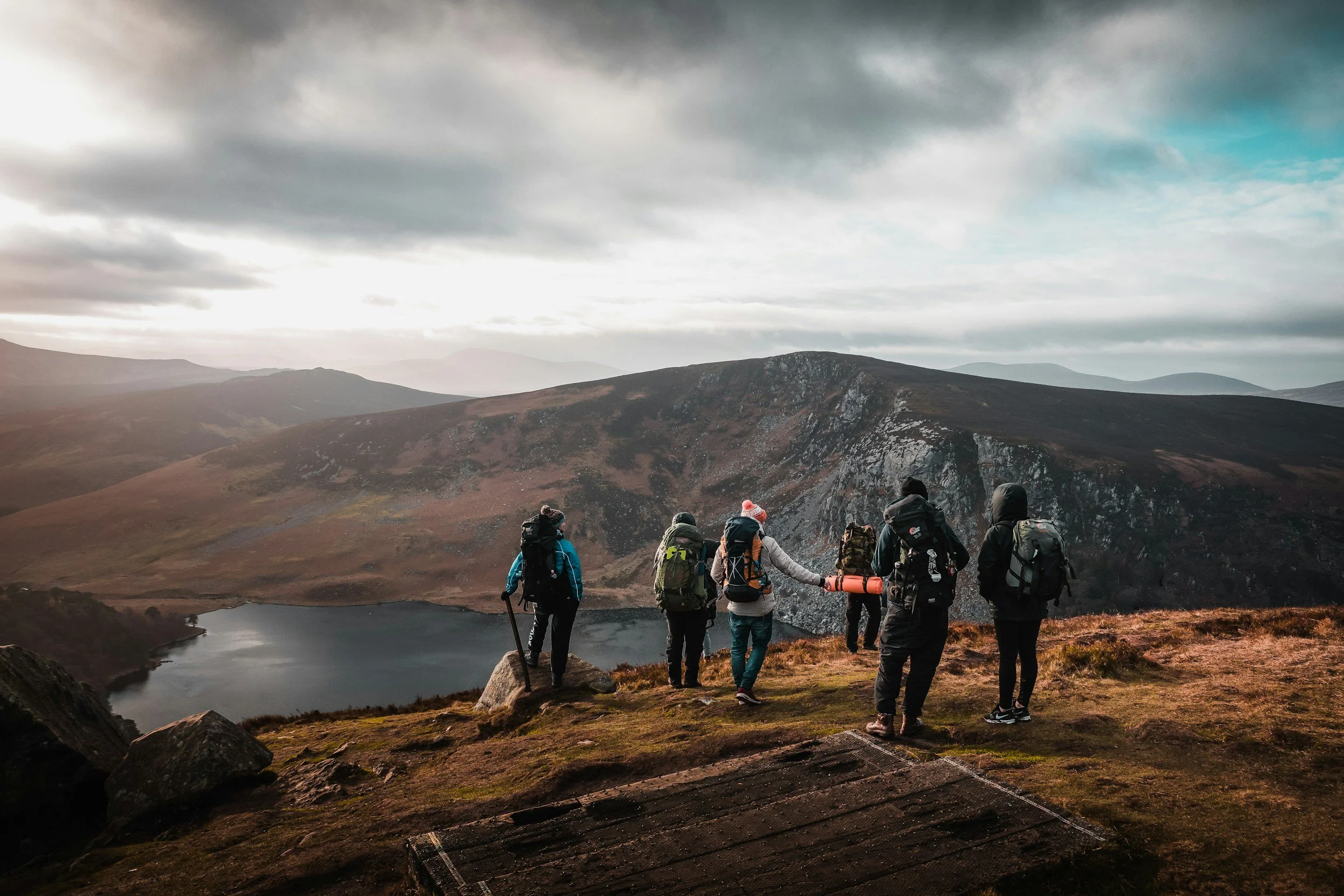 A group of hikers standing on a mountain trail, overlooking a mountain lake, with rolling hills and cloudy sky in the background.