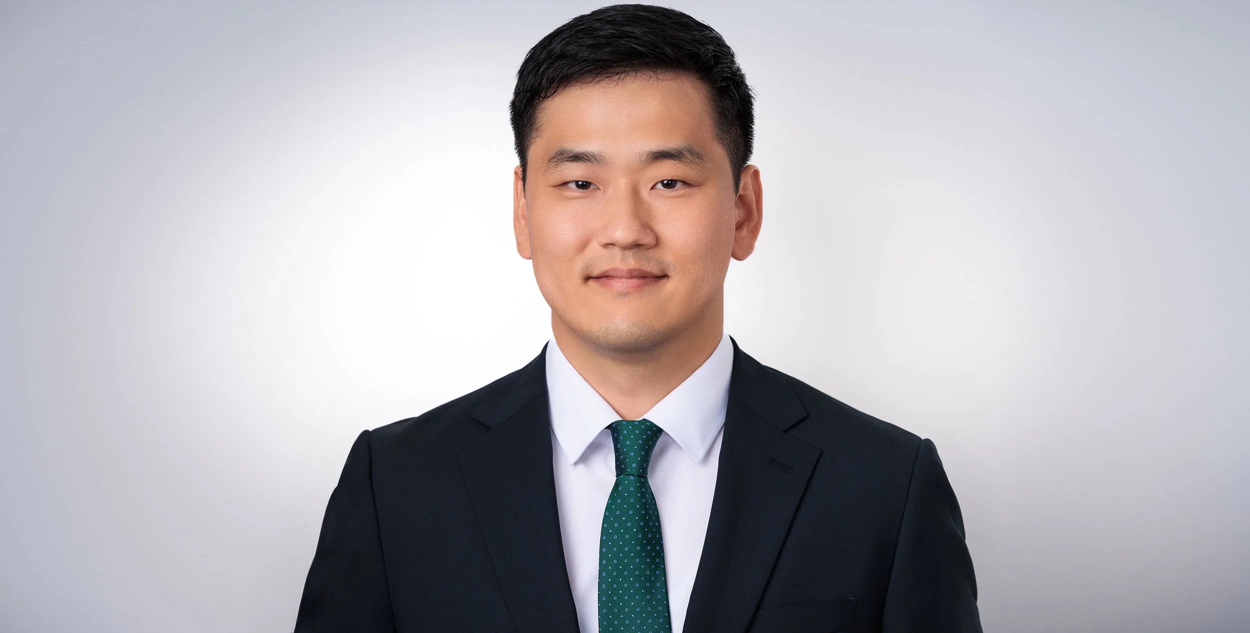 Professional headshot of a young Asian man in a suit and tie, smiling slightly against a plain light grey background.
